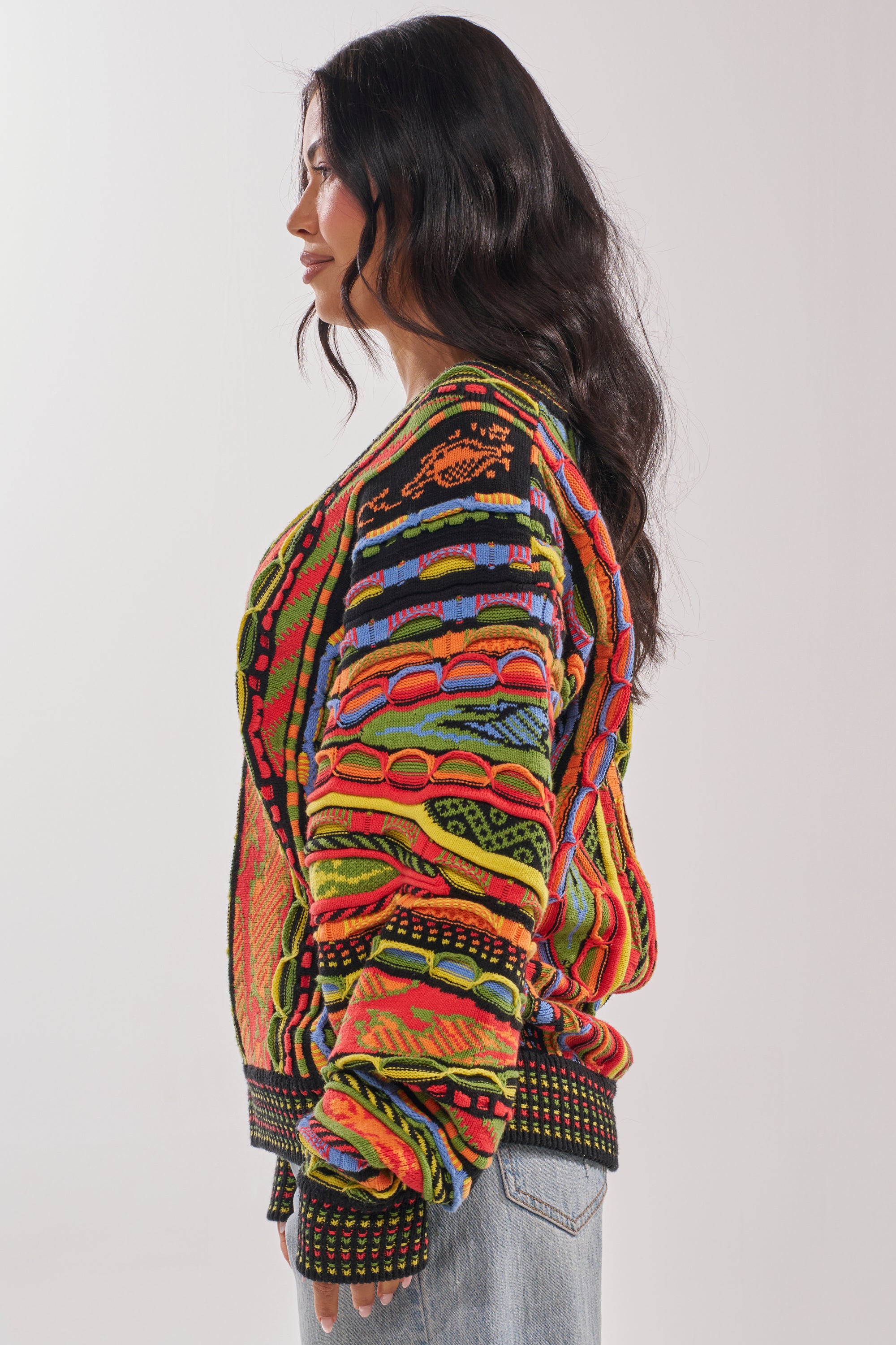 A person with long, wavy dark hair stands in profile, wearing the COSMIC WEAVE V-NECK SWEATER with geometric patterns and light blue jeans against a plain light background. 2