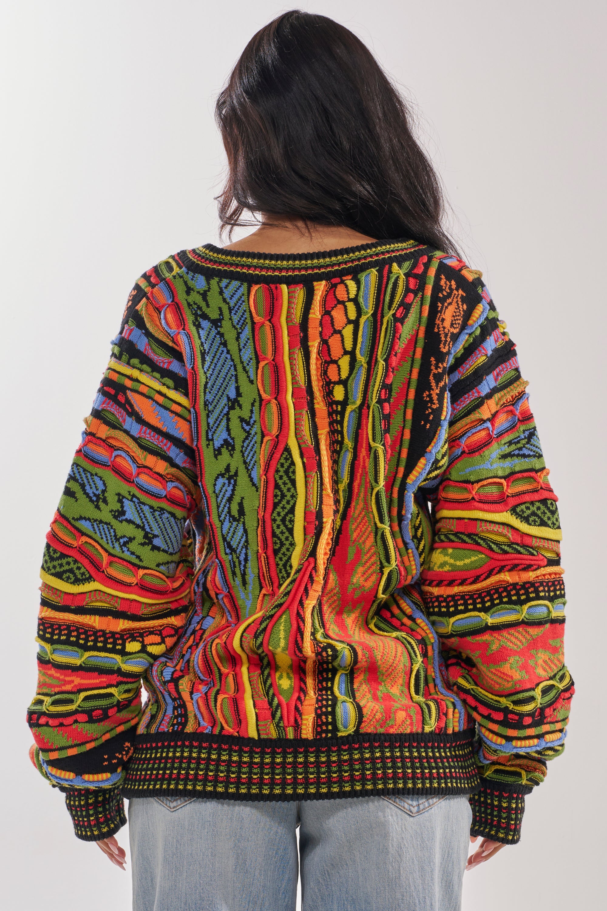 A person with long dark hair faces away from the camera, wearing the oversized COSMIC WEAVE V-NECK SWEATER with multicolor geometric and abstract designs in red, blue, yellow, green, and black. Light background. 3