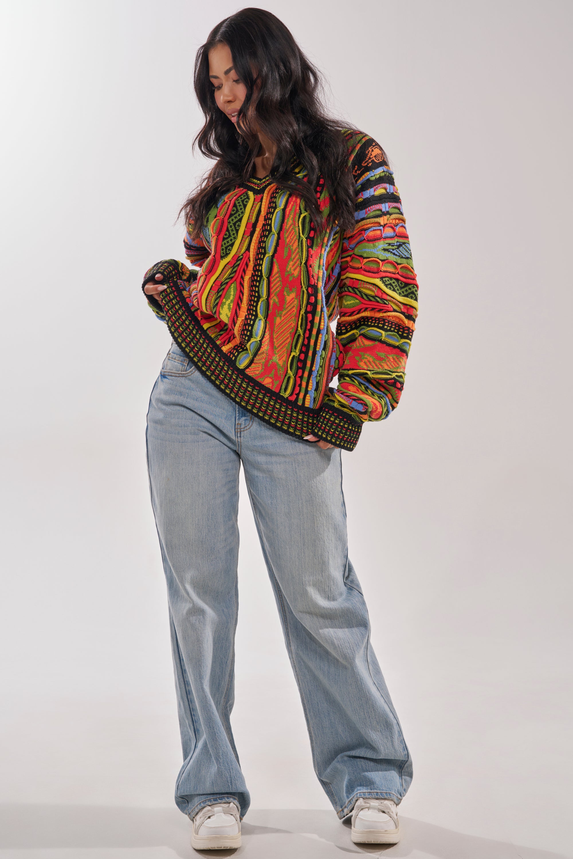 A woman with long dark hair wears the COSMIC WEAVE V-NECK SWEATER, styled with light blue wide-leg jeans and white sneakers, standing against a plain light background and looking downwards. 4