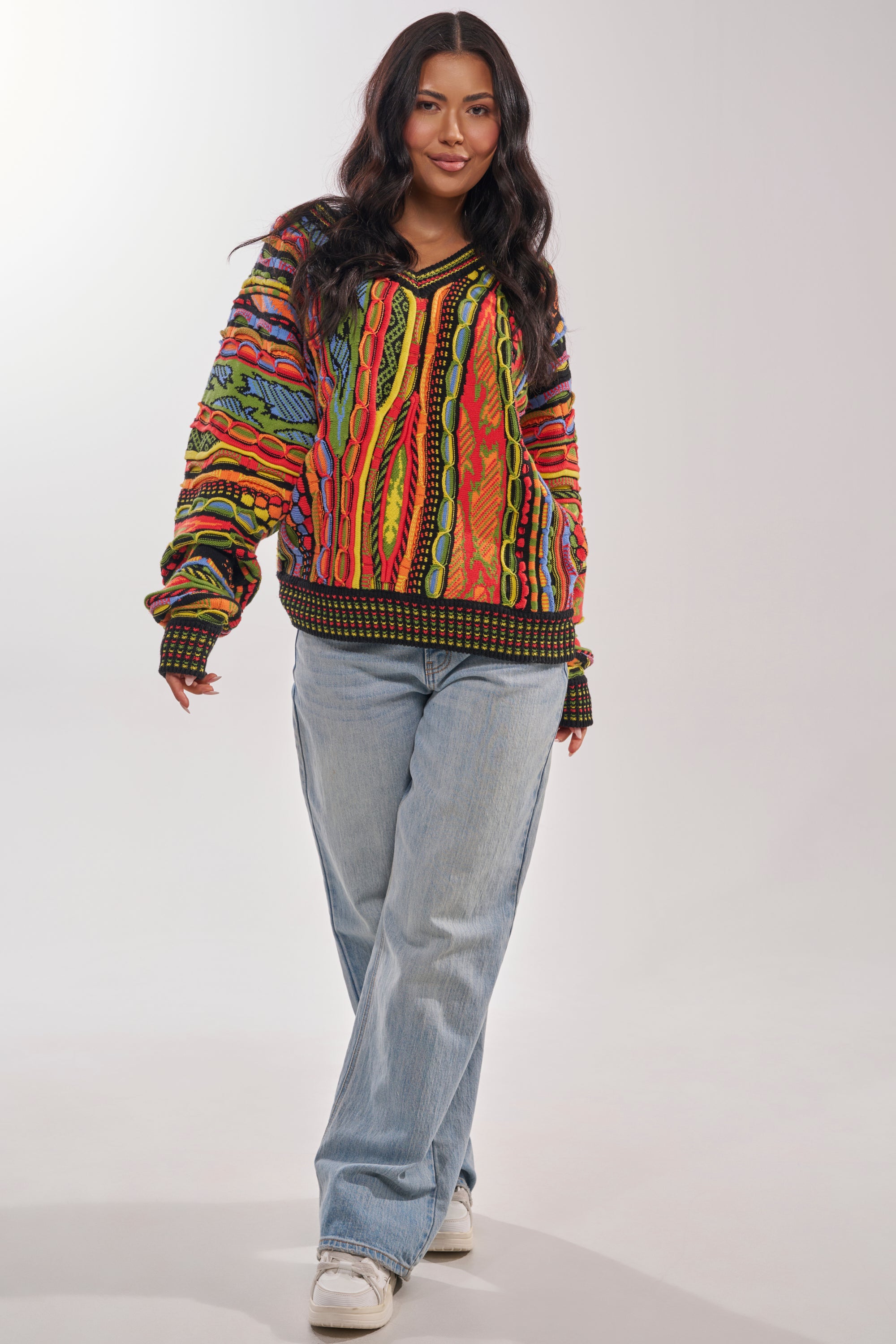 A woman with long dark hair wears the COSMIC WEAVE V-NECK SWEATER, paired with light blue jeans and white sneakers, standing and smiling against a plain light background. 5