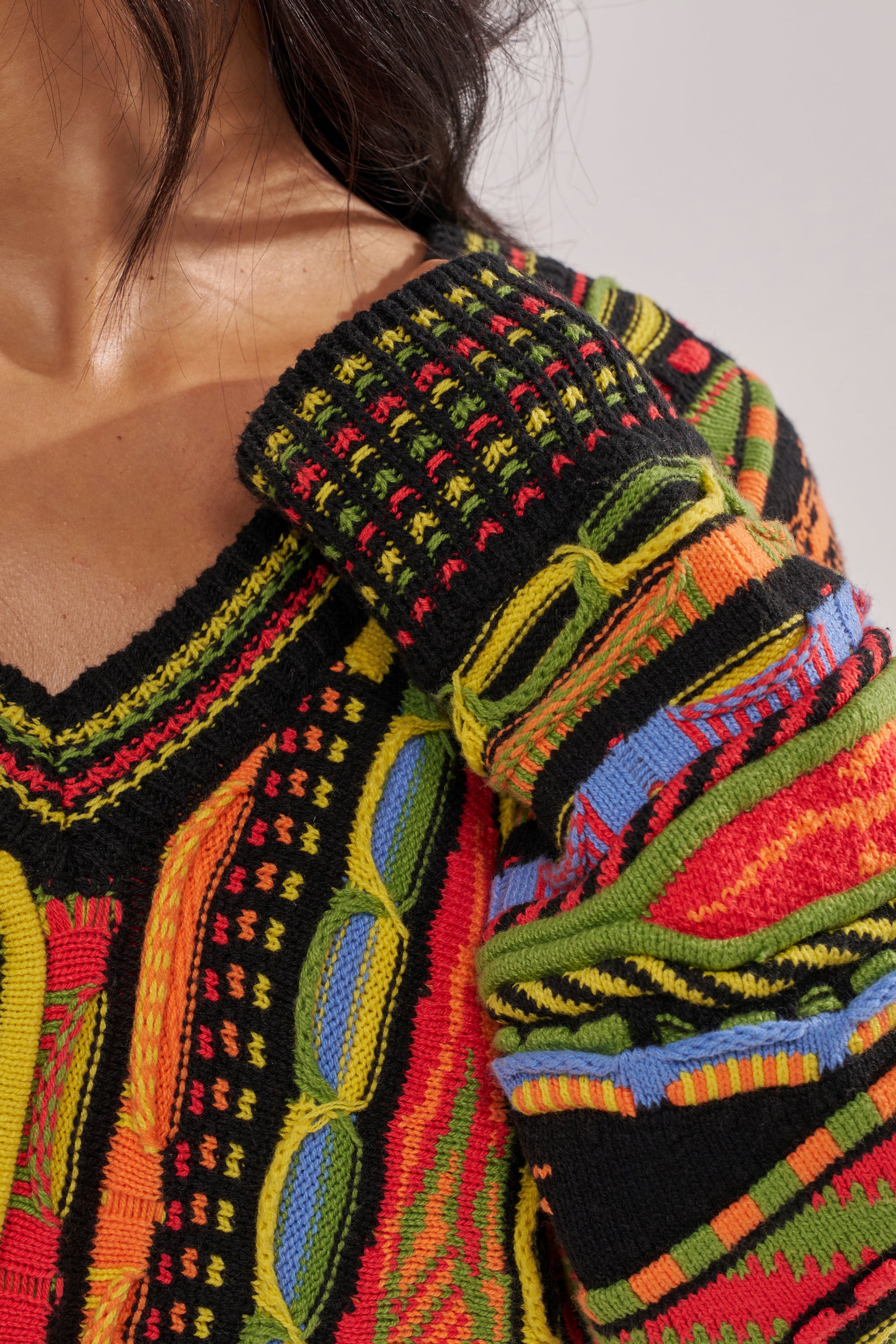 A close-up of a person wearing the COSMIC WEAVE V-NECK SWEATER—a black base sweater featuring bold yellow, red, blue, and green patterns. Their hand is resting on their shoulder. 6