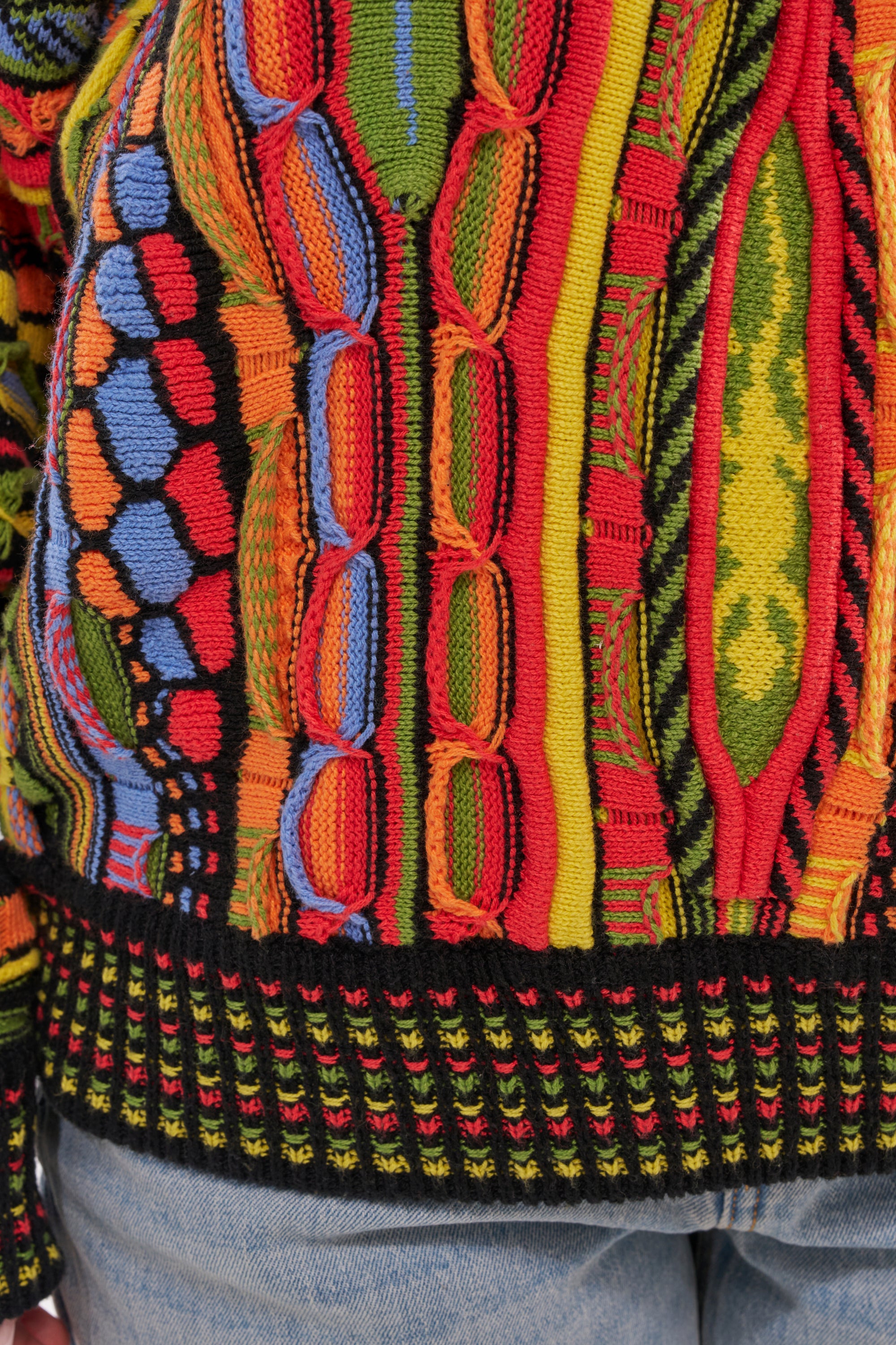 A close-up of the COSMIC WEAVE V-NECK SWEATER, featuring vibrant multicolor woven patterns in red, yellow, green, orange, and blue, paired with light blue jeans. 7