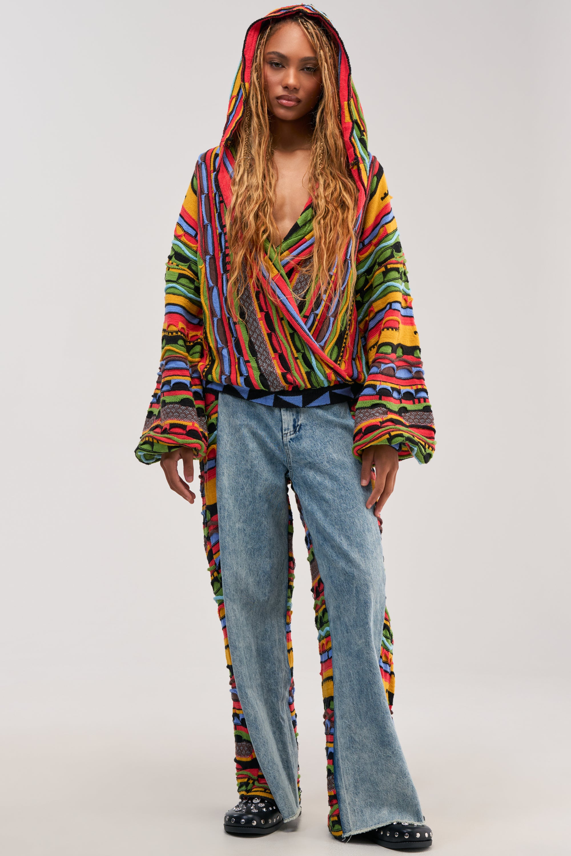 Wearing the ONE OF ONE DENIM WITH KNIT SIDE PANELS—blue wide leg jeans accented with unique knit side details—a person with long braids pairs them with a vibrant, hooded, striped top featuring multicolor knit accents and flared sleeves, and black studded shoes. Standing against a plain light background, this look makes a bold streetwear statement.