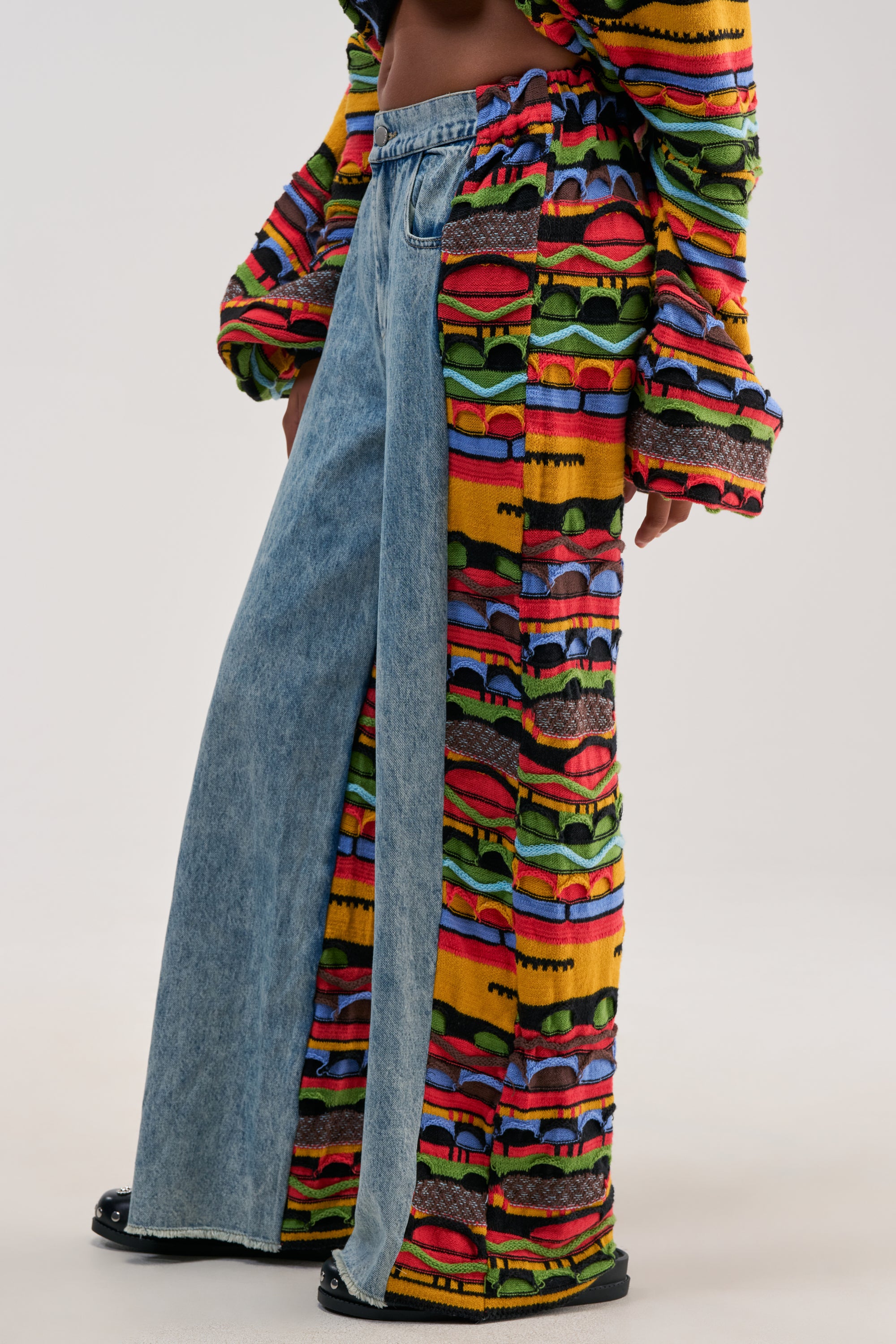 Wearing the ONE OF ONE DENIM WITH KNIT SIDE PANELS, this person makes a statement in streetwear with wide leg jeans—one side in classic blue denim and the other featuring colorful, patterned knit panels. They complete the look with a matching patterned top, leaving part of their midriff visible.