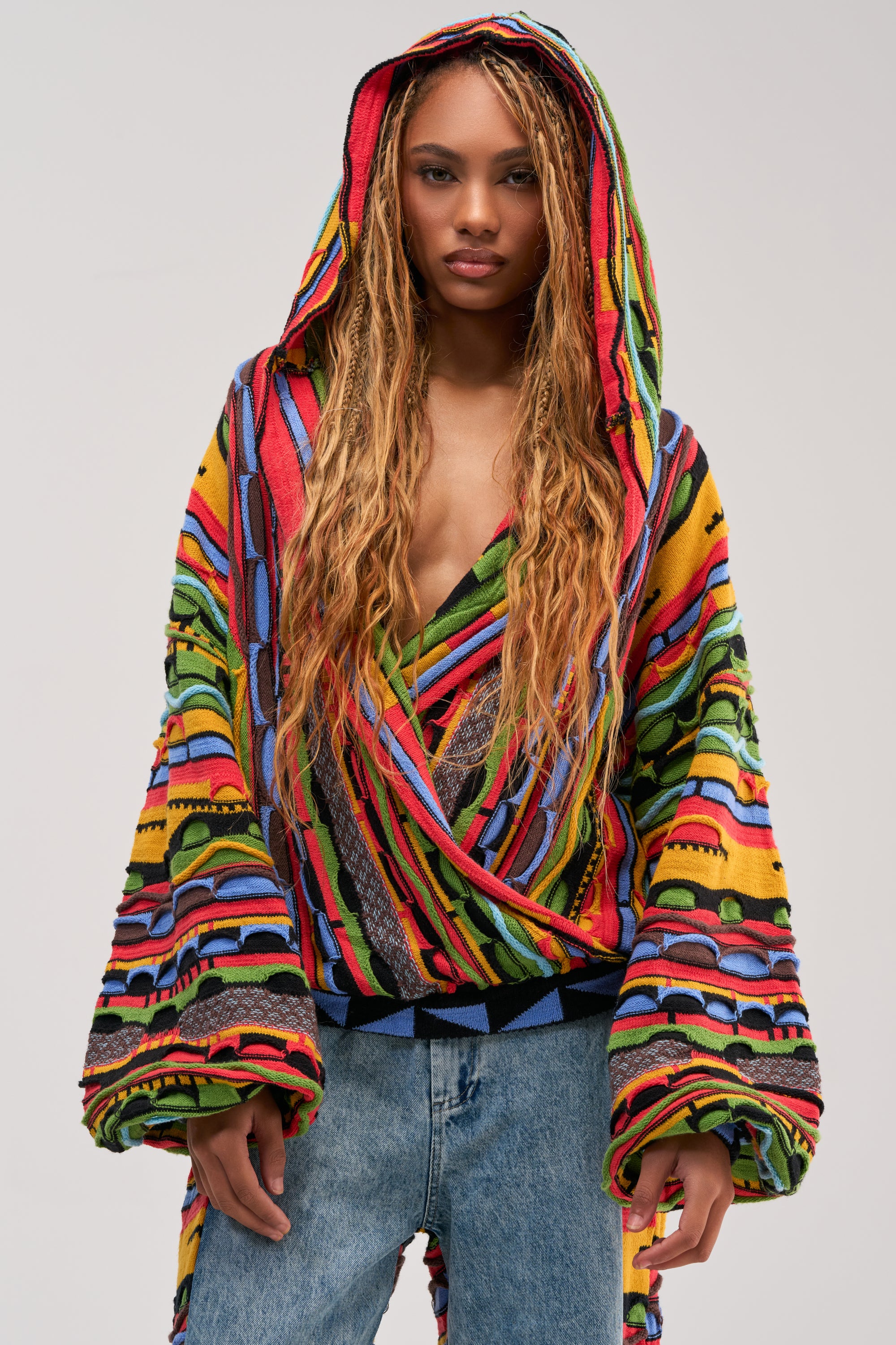A woman with long, braided hair models the ONE OF ONE WRAP SWEATER—a colorful striped sweater featuring a hood and wide sleeves—styled with blue jeans, against a plain light background.