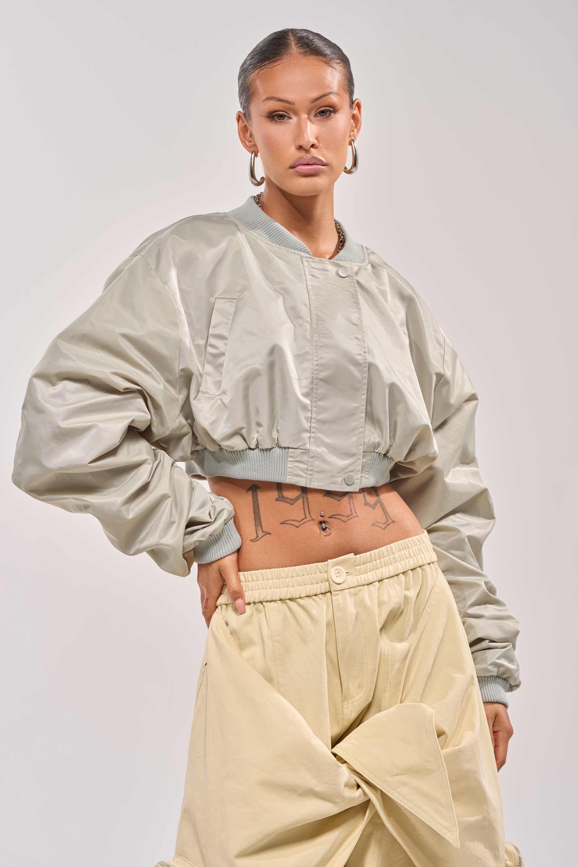 A person with slicked-back hair wears the TAKE IT EASY CROPPED BOMBER IN SAGE, beige wide-leg pants with a knotted front, large hoop earrings, and showcases visible tattoos on their midriff while posing confidently against a plain background. 0