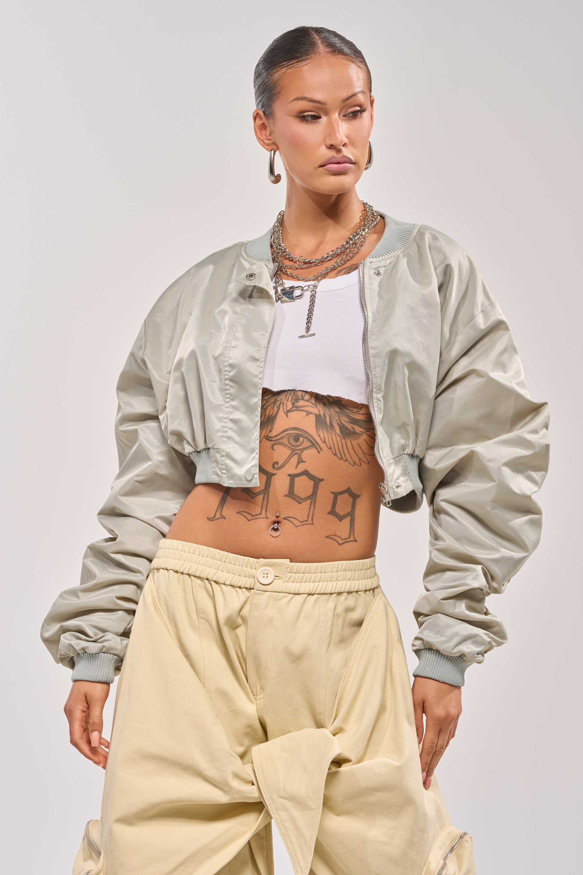 A woman with slicked-back hair wears the TAKE IT EASY CROPPED BOMBER IN SAGE over a white crop top, beige baggy pants, hoop earrings, and layered necklaces. A large tattoo is visible across her stomach. 2