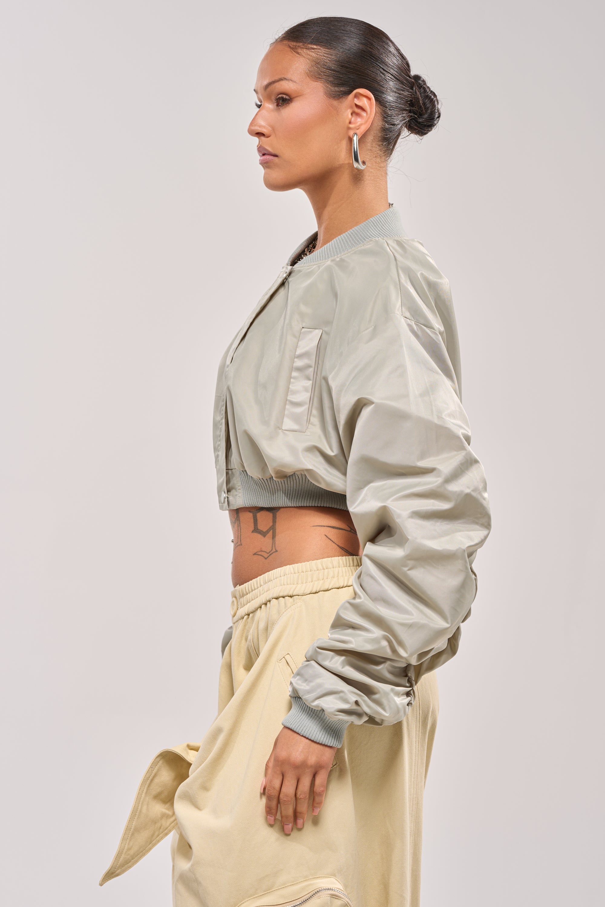 A woman with slicked-back hair wears the TAKE IT EASY CROPPED BOMBER IN SAGE, beige pants, and silver hoop earrings. Standing in profile against a plain background, she reveals a tattoo on her waist. 3