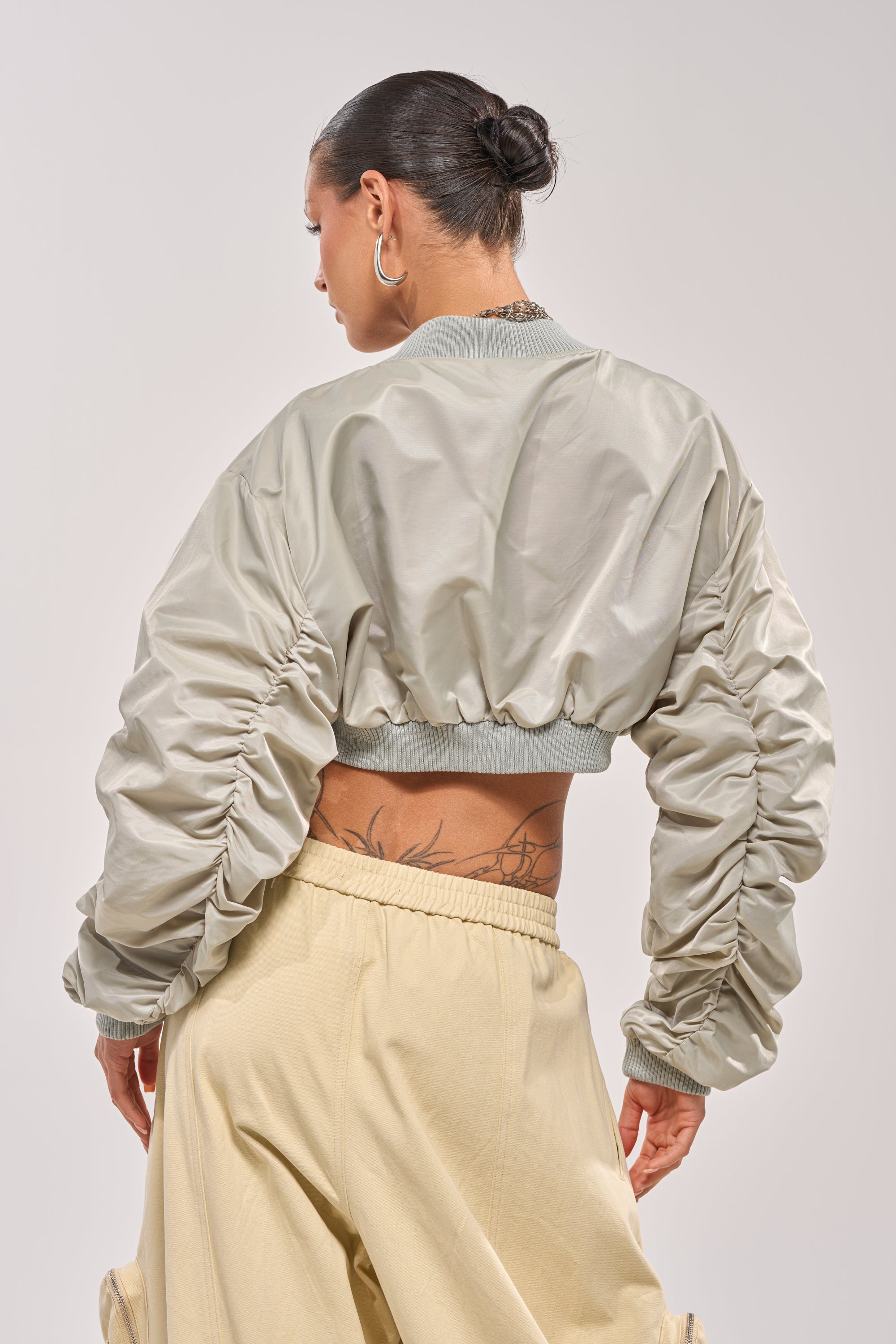 A person faces away, wearing the TAKE IT EASY CROPPED BOMBER IN SAGE with ruched sleeves and loose beige pants. Their hair is in a bun, revealing a lower back tattoo—an effortlessly cool courtside bomber look. 4
