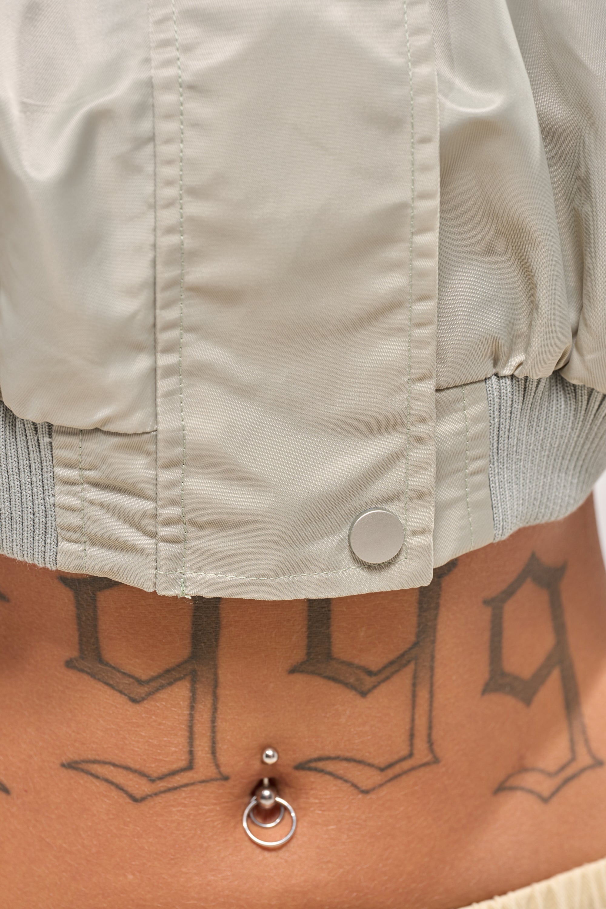 Close-up of a person's lower torso with a metallic belly button ring and "1999" tattoo in bold, styled effortlessly with the TAKE IT EASY CROPPED BOMBER IN SAGE for a chic courtside look. 6
