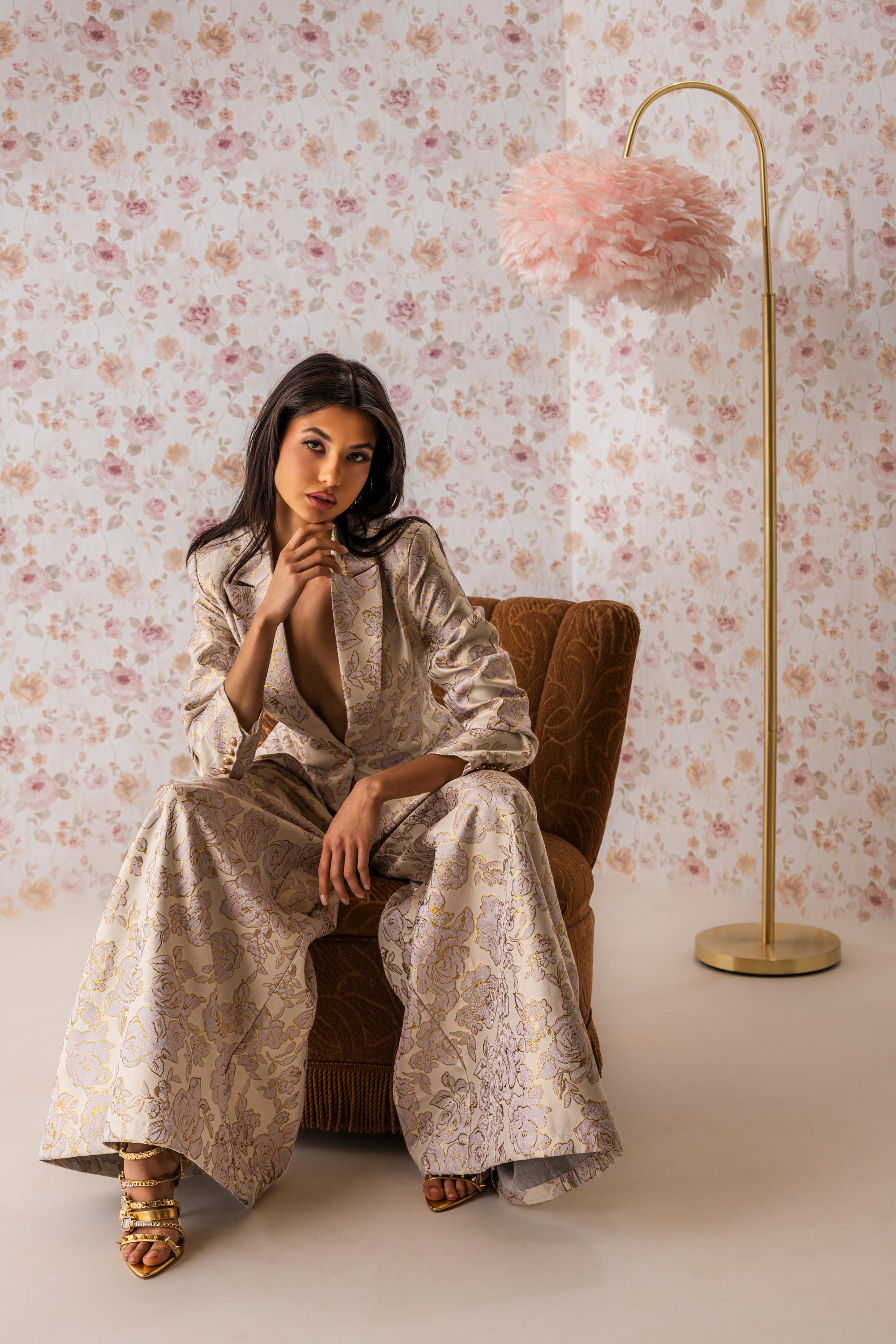 A woman wearing the CHERI BROCADE WIDE LEG PANT sits on a brown chair in a floral-walled room, resting her arm on her knee and gazing at the camera beside a gold floor lamp topped with a pink feather lampshade. 0