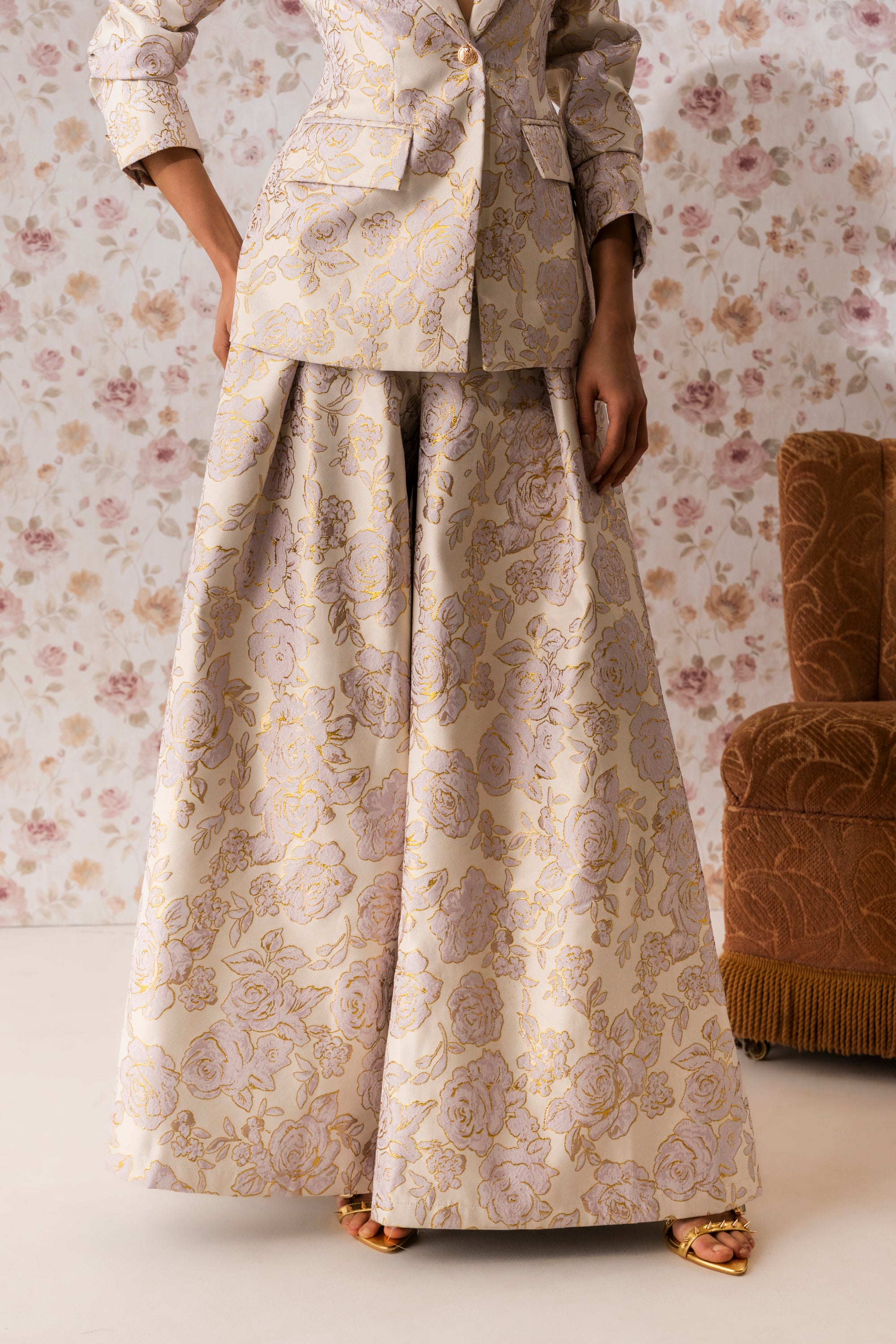 A person stands in a room wearing the CHERI BROCADE WIDE LEG PANT with a high-rise waist. Only their lower half is visible, while floral wallpaper and a brown chair are seen in the background. 1