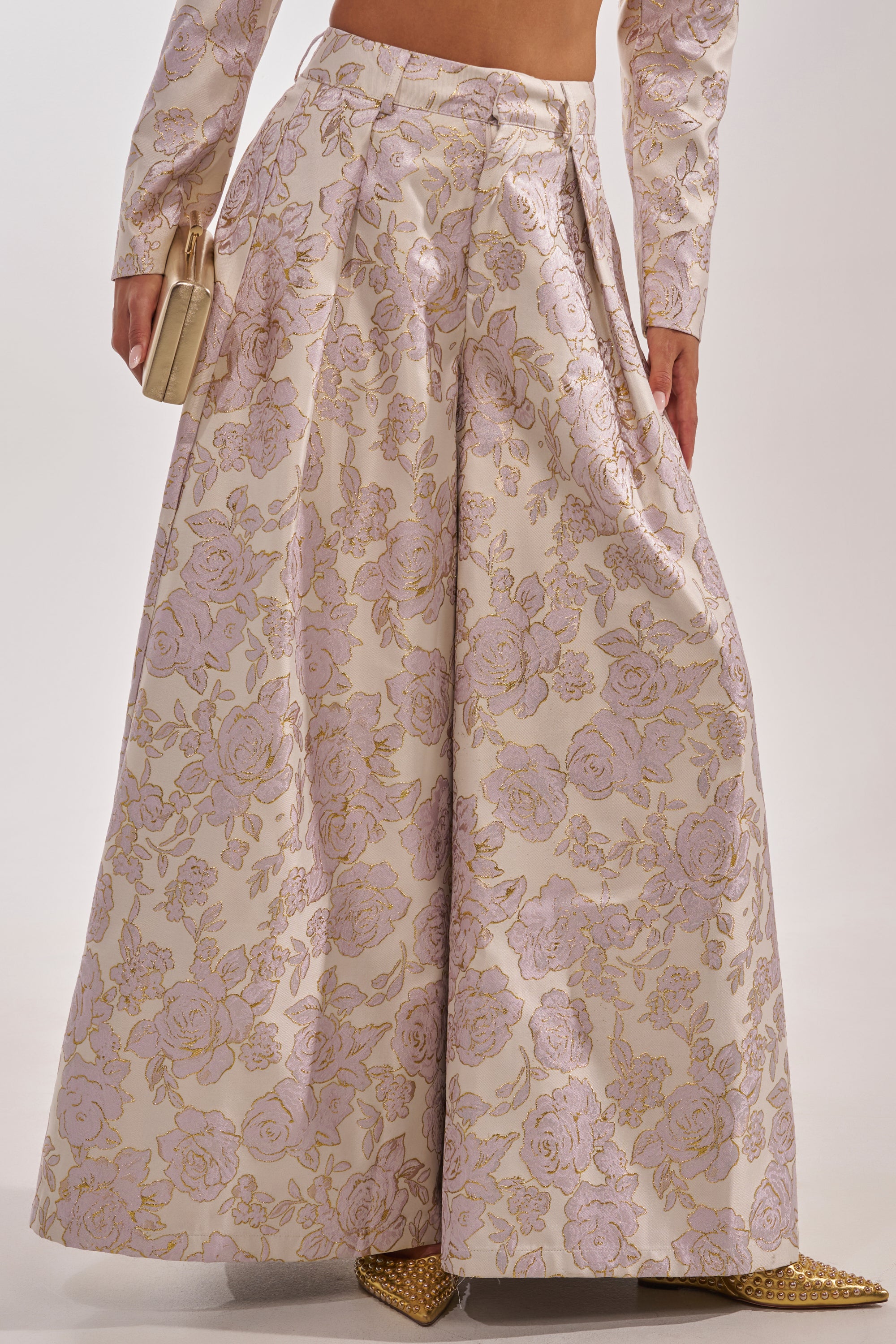 A person wearing CHERI BROCADE WIDE LEG PANT in a pale pink and gold floral pattern, paired with a matching cropped top, holding a small beige purse, and gold pointed shoes. 4