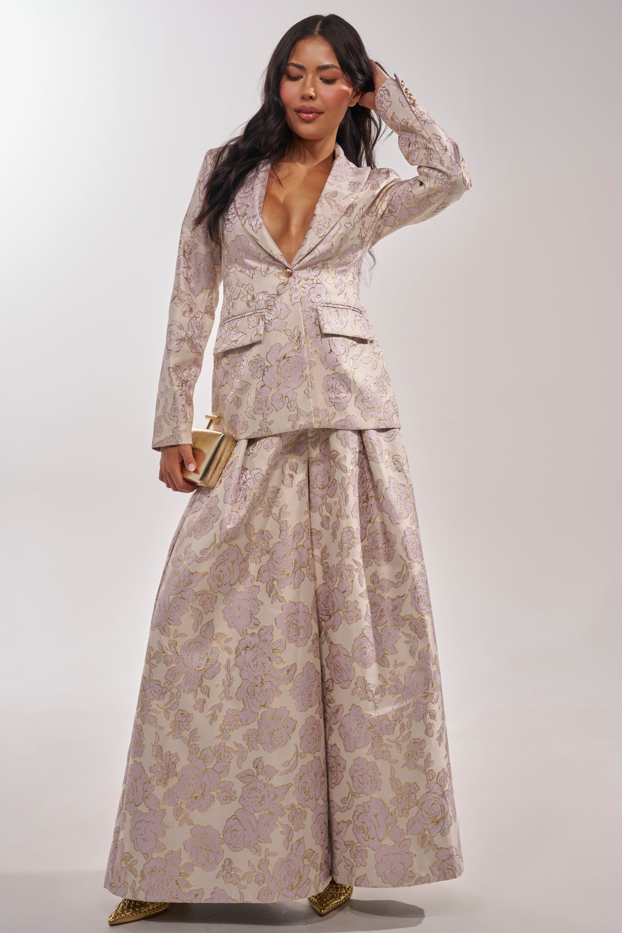 A woman models the CHERI BROCADE WIDE LEG PANT with a floral blazer featuring a deep v-neck. She holds a gold clutch, wears pointed gold shoes, and her long dark hair flows in loose waves against a plain light background. 7