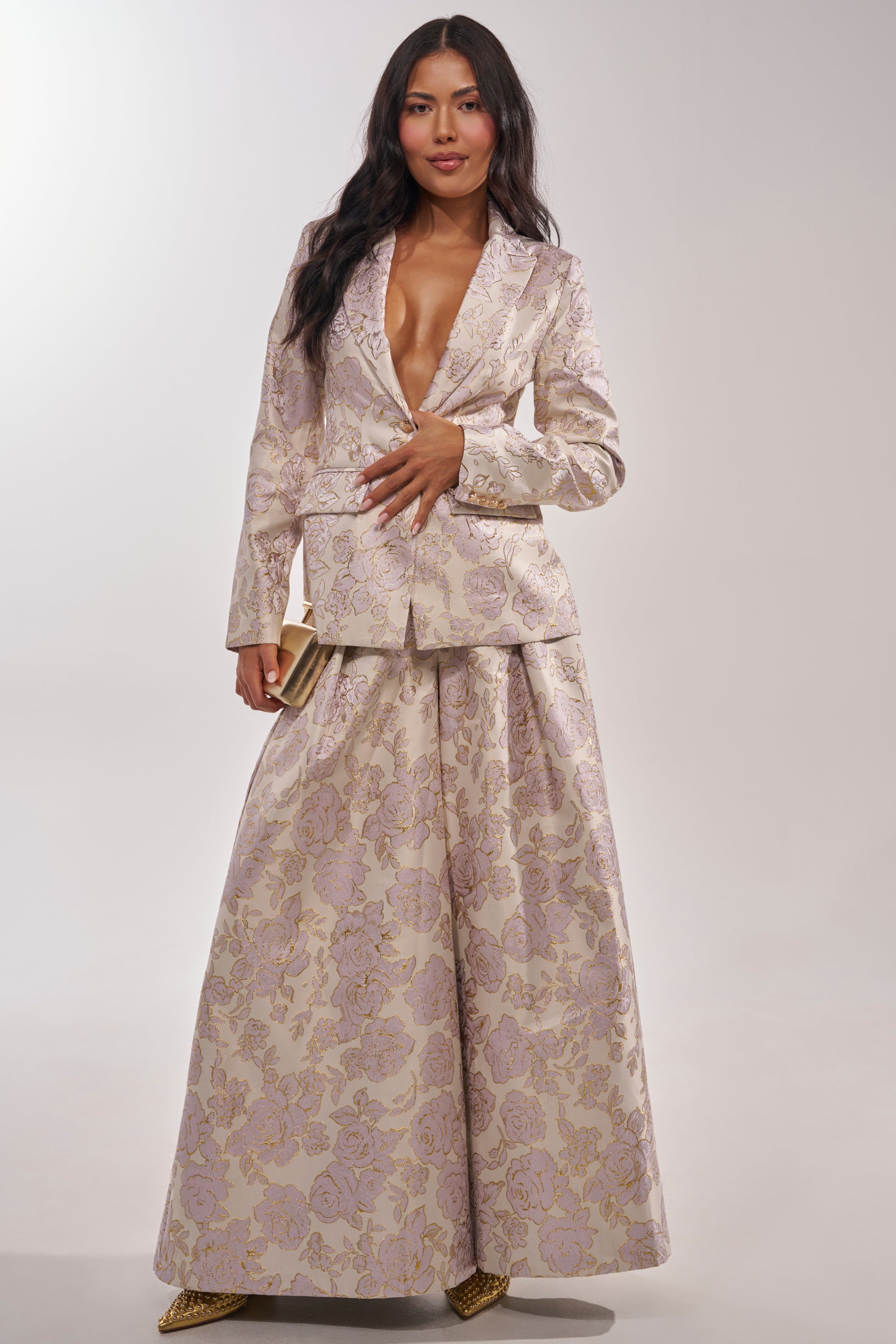 A woman poses confidently in CHERI BROCADE WIDE LEG PANT, featuring metallic floral details and a high-rise waist, styled with a deep V-neck blazer, gold pointed shoes, long dark hair, and a clutch against a plain background. 8