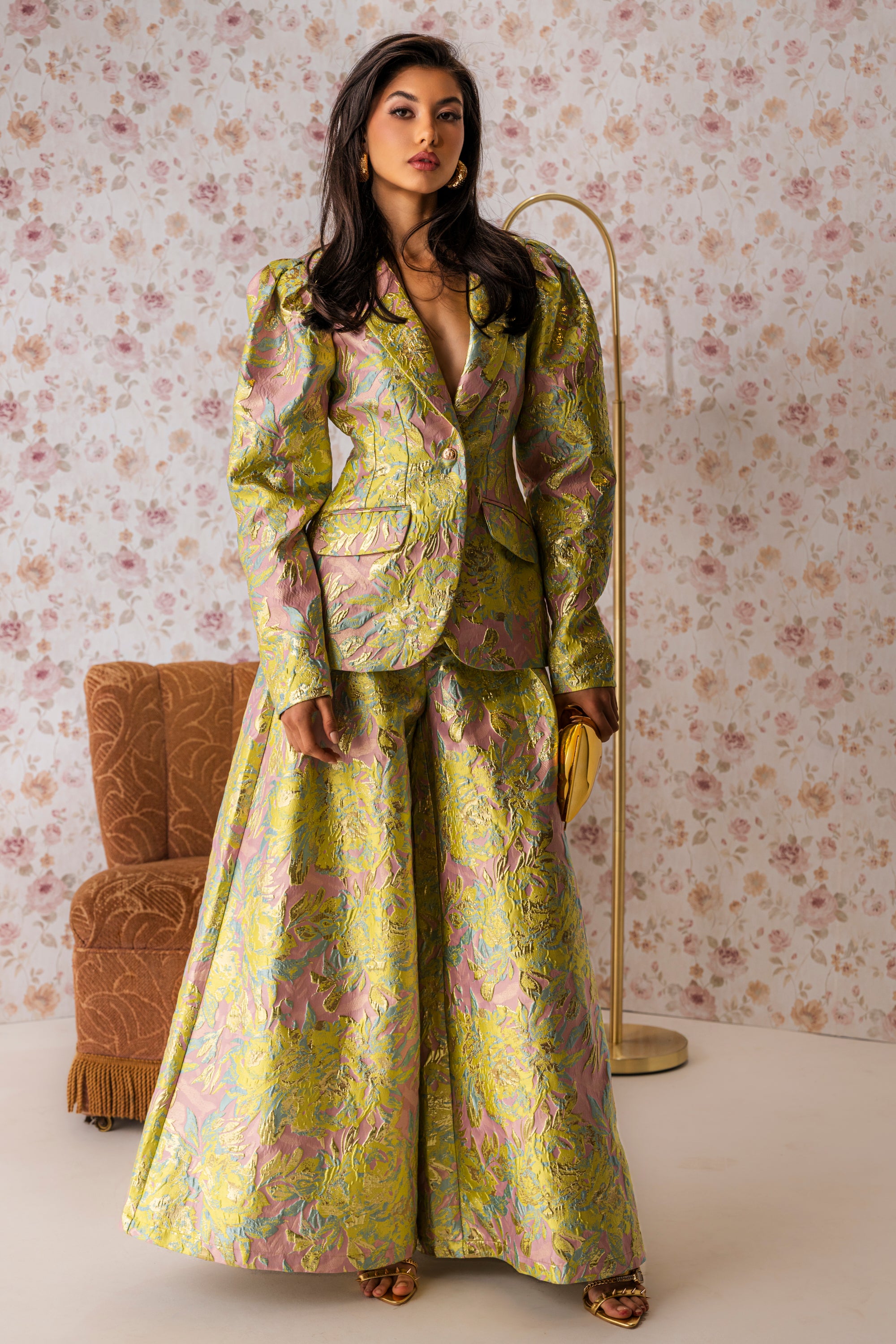 A woman stands indoors wearing the LUCY BROCADE WIDE LEG PANT, a shiny suit with ultra-wide leg trousers and an iridescent floral pattern. She holds a small clutch and gold heels, set against floral wallpaper and stylish décor. 0