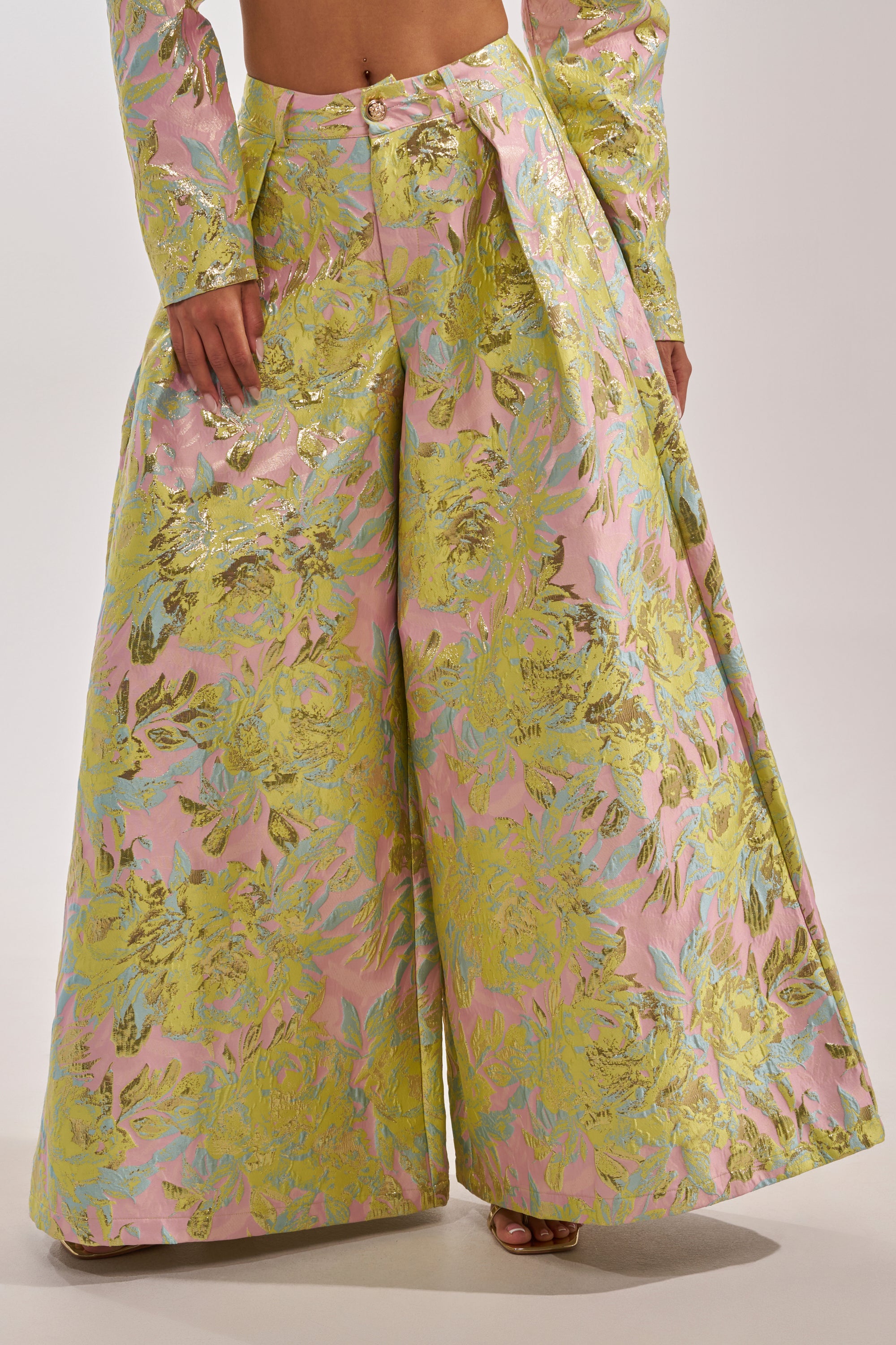 A person wears the LUCY BROCADE WIDE LEG PANT, featuring an ultra-wide leg and a bold metallic floral pattern in yellow, pink, and green, paired with a matching long-sleeve top for a dramatic silhouette. 3