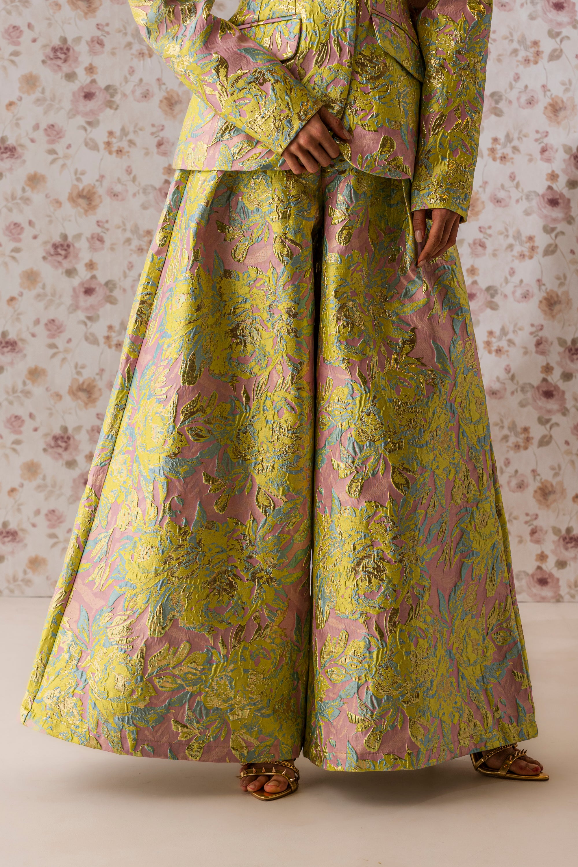 Wearing the LUCY BROCADE WIDE LEG PANT, a person stands against floral wallpaper. Their pastel pink and green jacket and iridescent floral trousers reflect the pattern, with only the lower face seen above open-toed sandals. 1