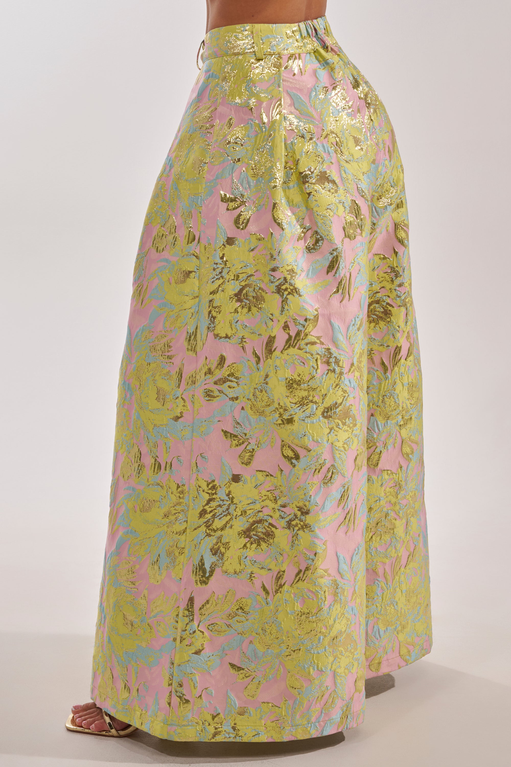 Someone wearing the LUCY BROCADE WIDE LEG PANT, featuring metallic wide-leg trousers with a shiny yellow-green and gold floral pattern on a light pink background, styled with open-toe heels. 4