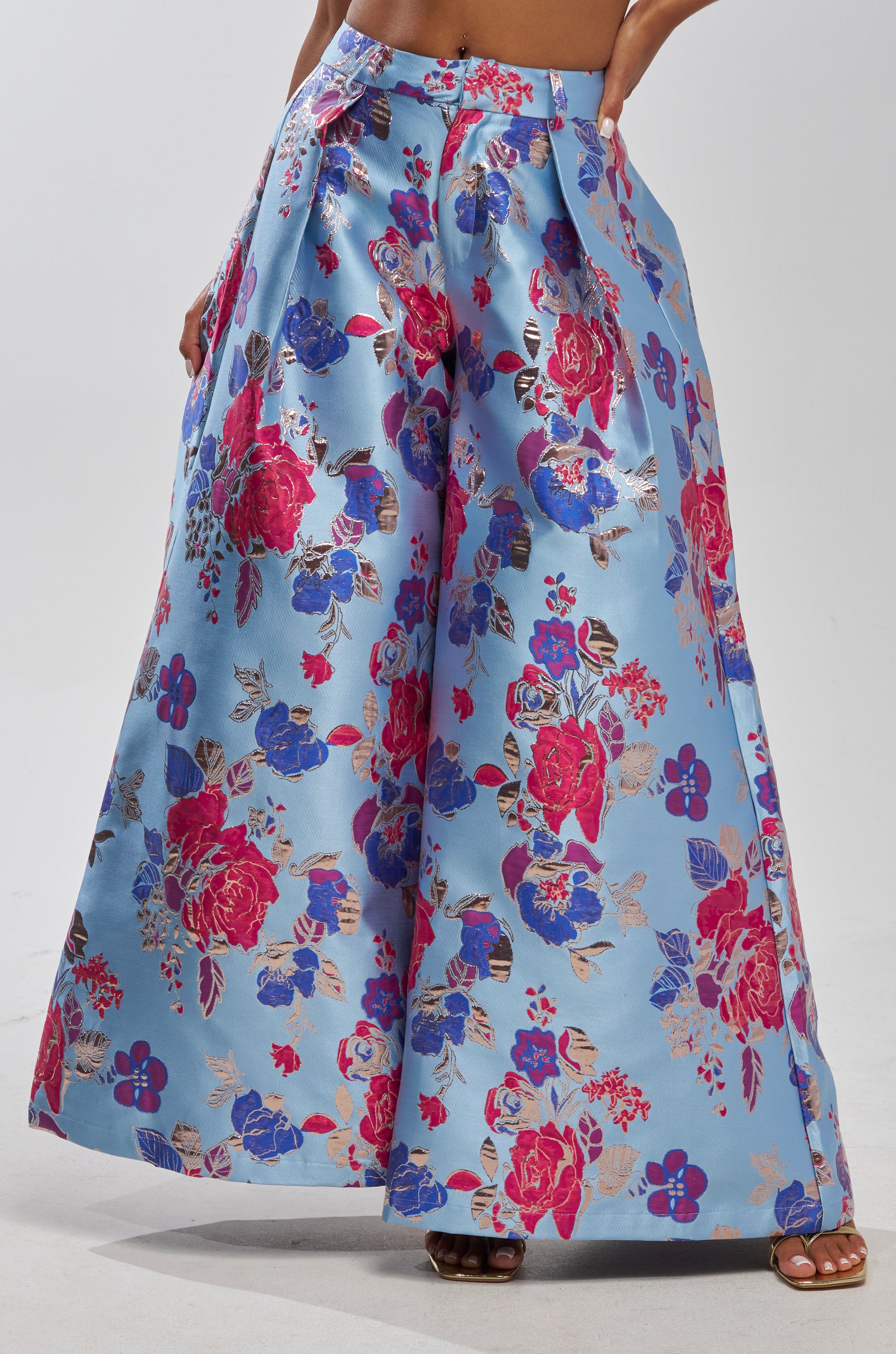 A person wears the BUY MYSELF FLOWERS BROCADE TROUSER IN BLUE, a high-waisted, wide-leg style with bold red, pink, and blue floral patterns on a light blue background. Features pockets and an elegant, flowing silhouette.