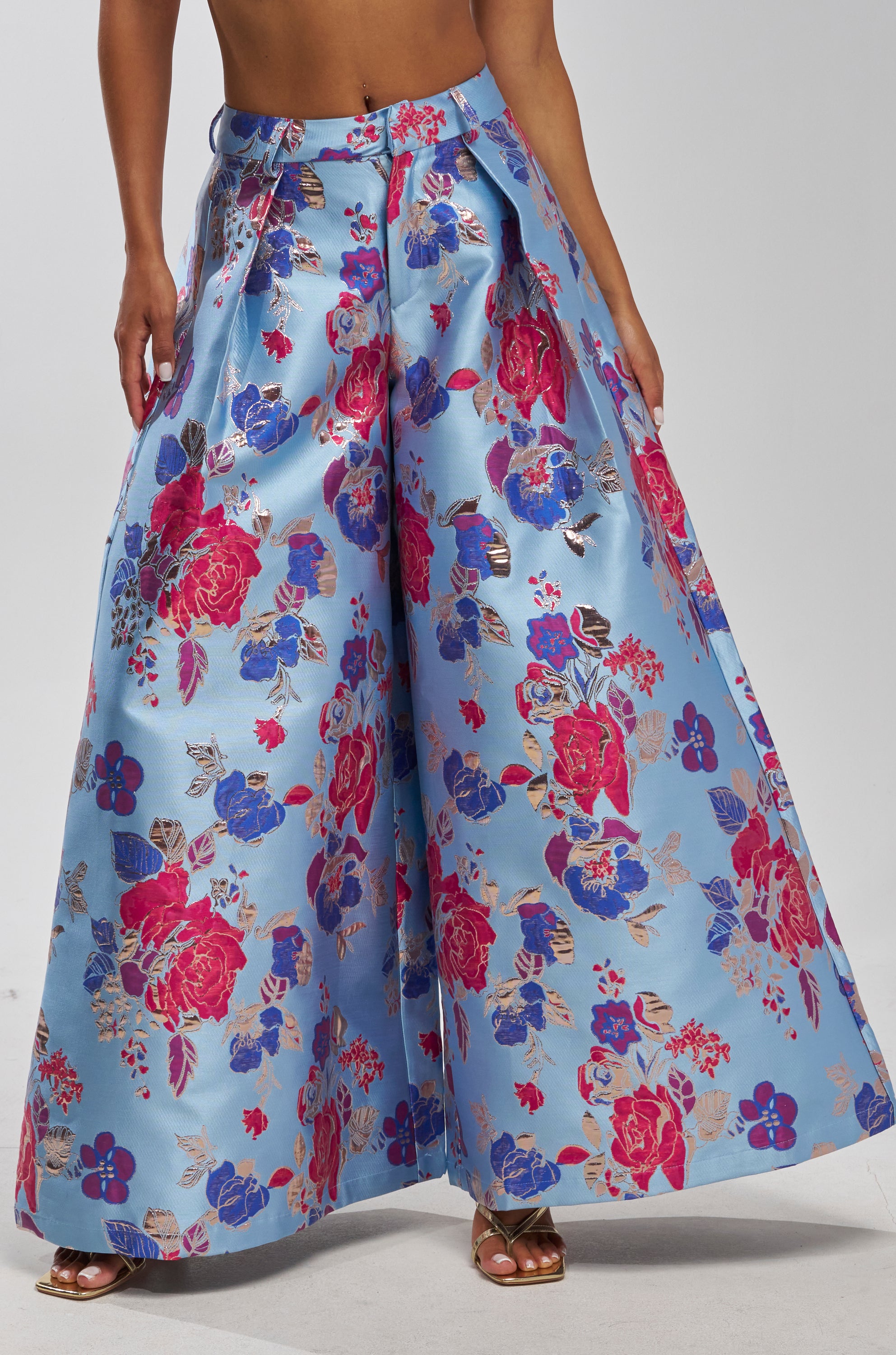 Someone models the BUY MYSELF FLOWERS BROCADE TROUSER IN BLUE—high-waisted, wide-leg pants featuring shiny light blue fabric with bold red, pink, and blue floral patterns. The wearer is standing with their face out of view.