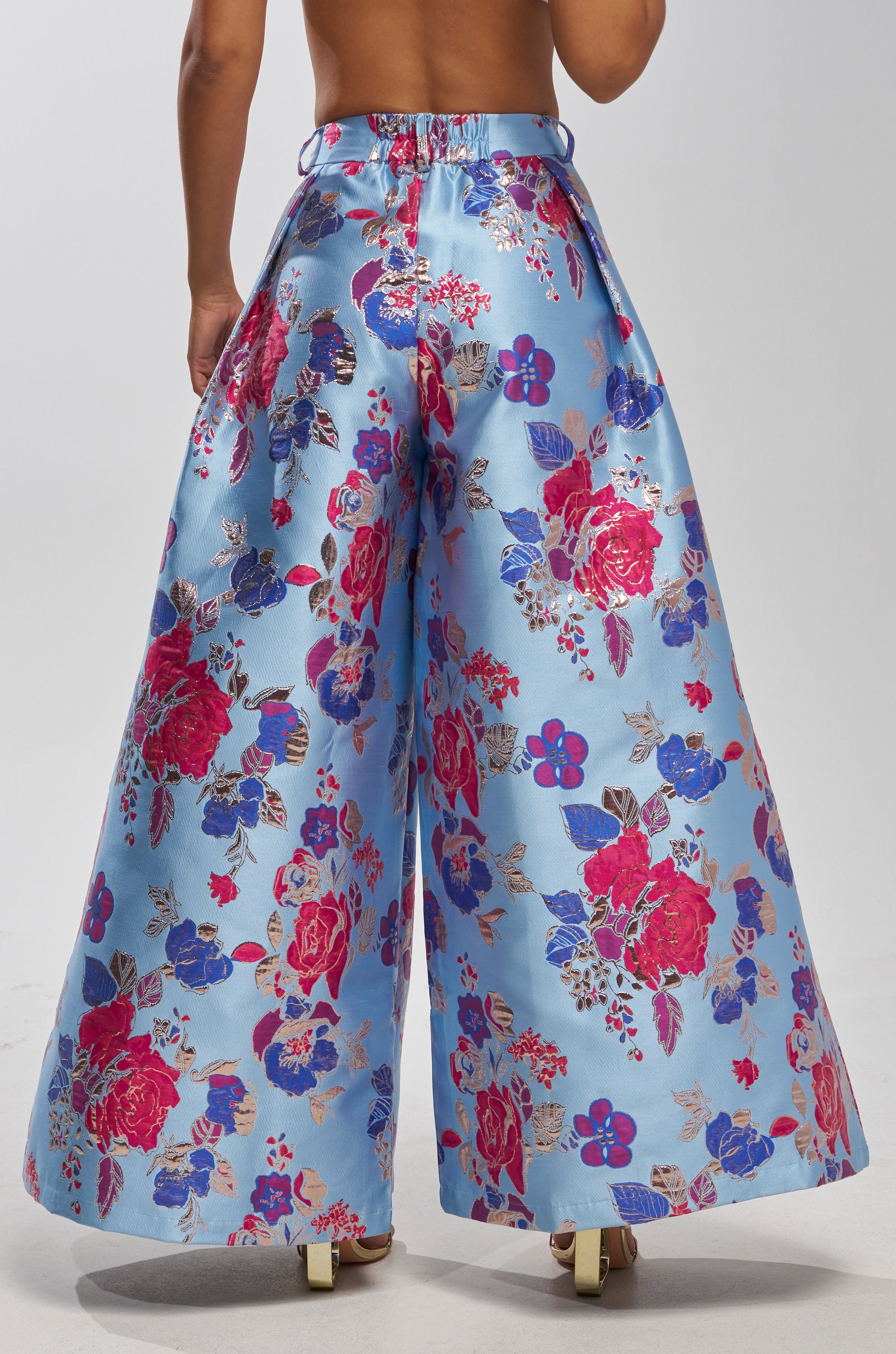 Someone wears the BUY MYSELF FLOWERS BROCADE TROUSER IN BLUE, a high-waisted wide-leg formal trouser featuring a shiny light blue background and vibrant floral print in red, pink, blue, and purple, standing against a plain white backdrop.