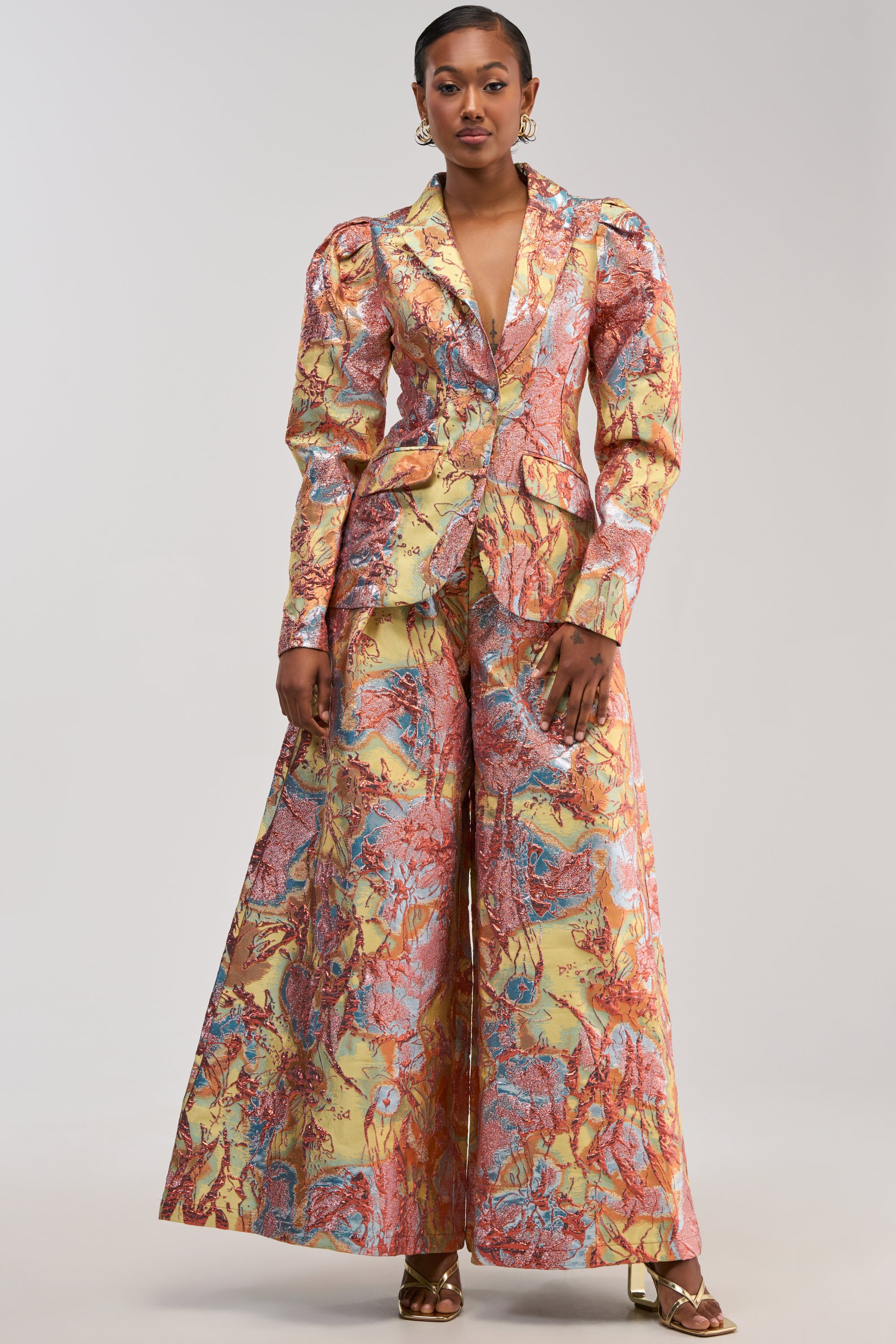 A woman poses against a plain light background, wearing the LUCY BROCADE WIDE LEG PANT IN BLUE MULTI paired with a fitted, colorful blazer. She completes her look with gold heels and jewelry.