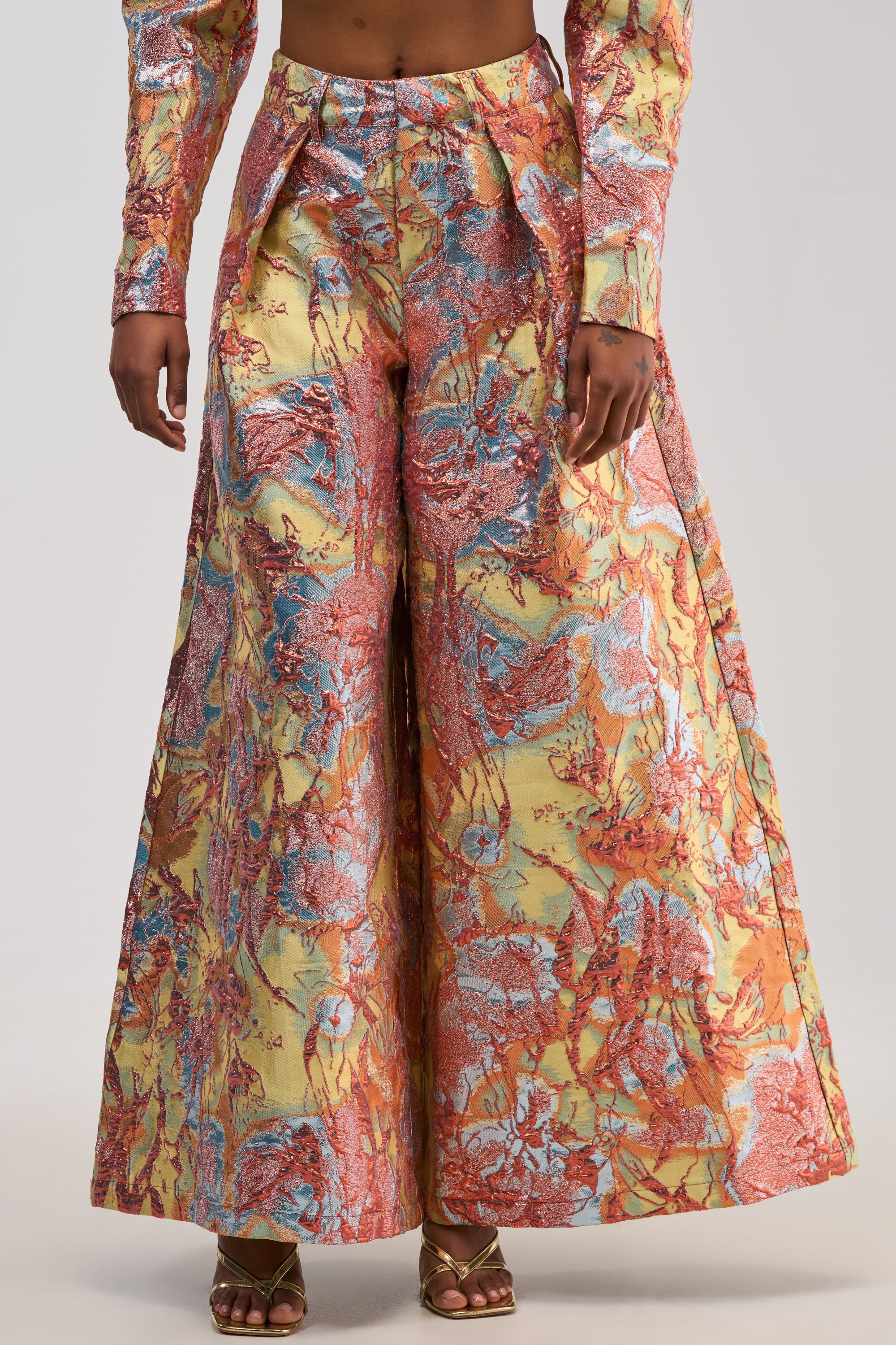 A person wears the LUCY BROCADE WIDE LEG PANT IN BLUE MULTI, featuring a colorful, abstract marble pattern in red, blue, and yellow, styled with a matching long-sleeve top and gold open-toe heels.