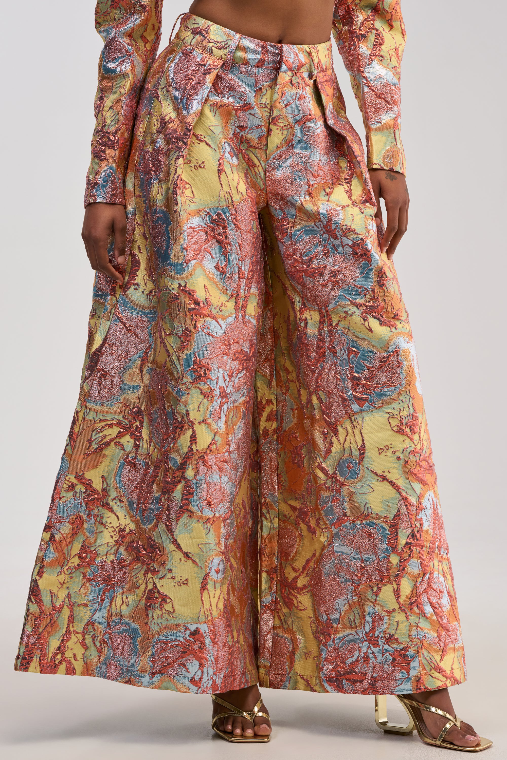 A person wearing the LUCY BROCADE WIDE LEG PANT IN BLUE MULTI, a high-waisted, wide-leg statement pant featuring a metallic abstract pattern in orange, yellow, blue, and gold, paired with matching long sleeves and gold heeled sandals.