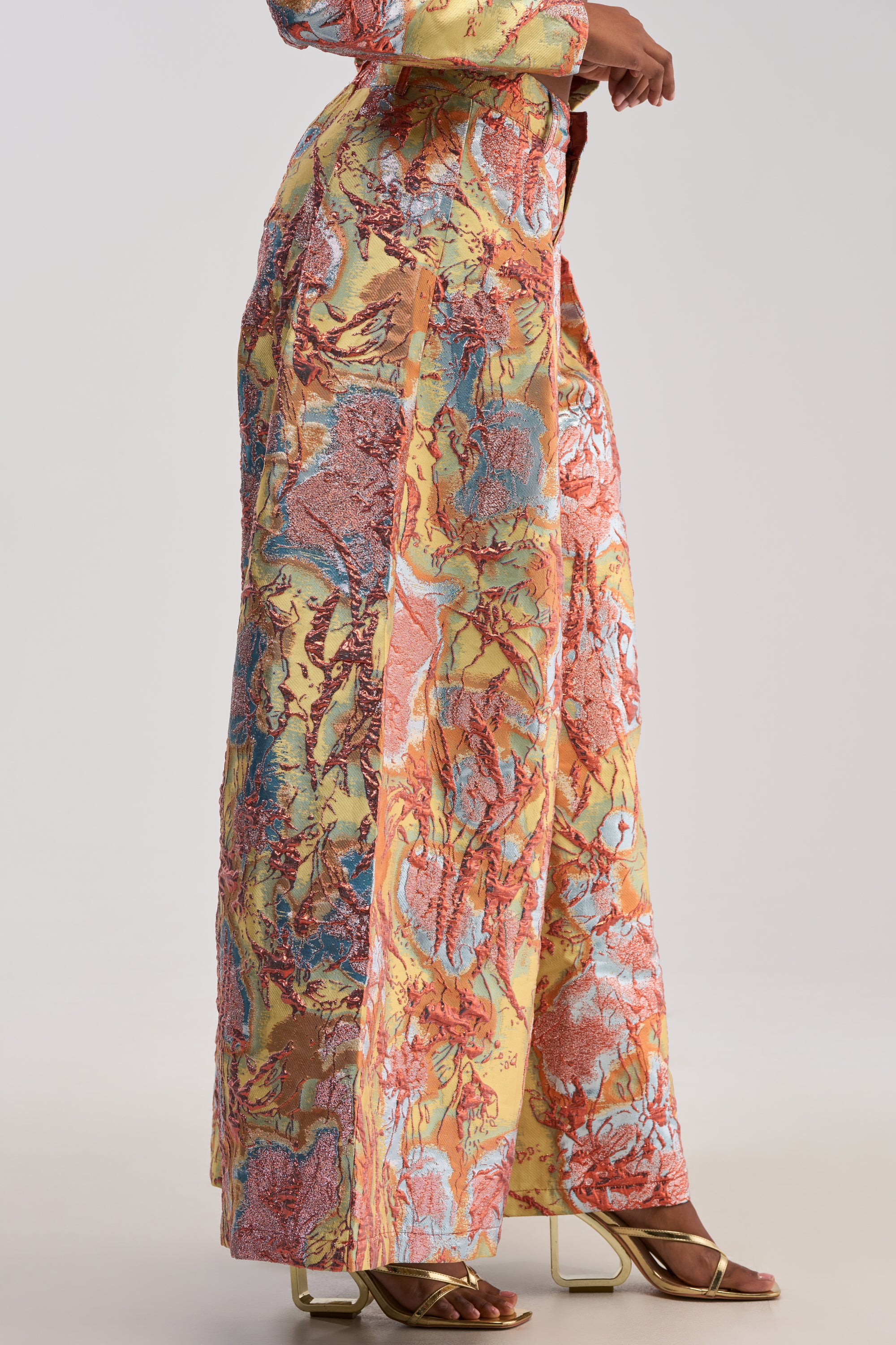 A person wearing the LUCY BROCADE WIDE LEG PANT IN BLUE MULTI, featuring a long, colorful abstract pattern in pink, yellow, and blue. Only the lower part of the body is visible, and the pants are paired with gold sandals.