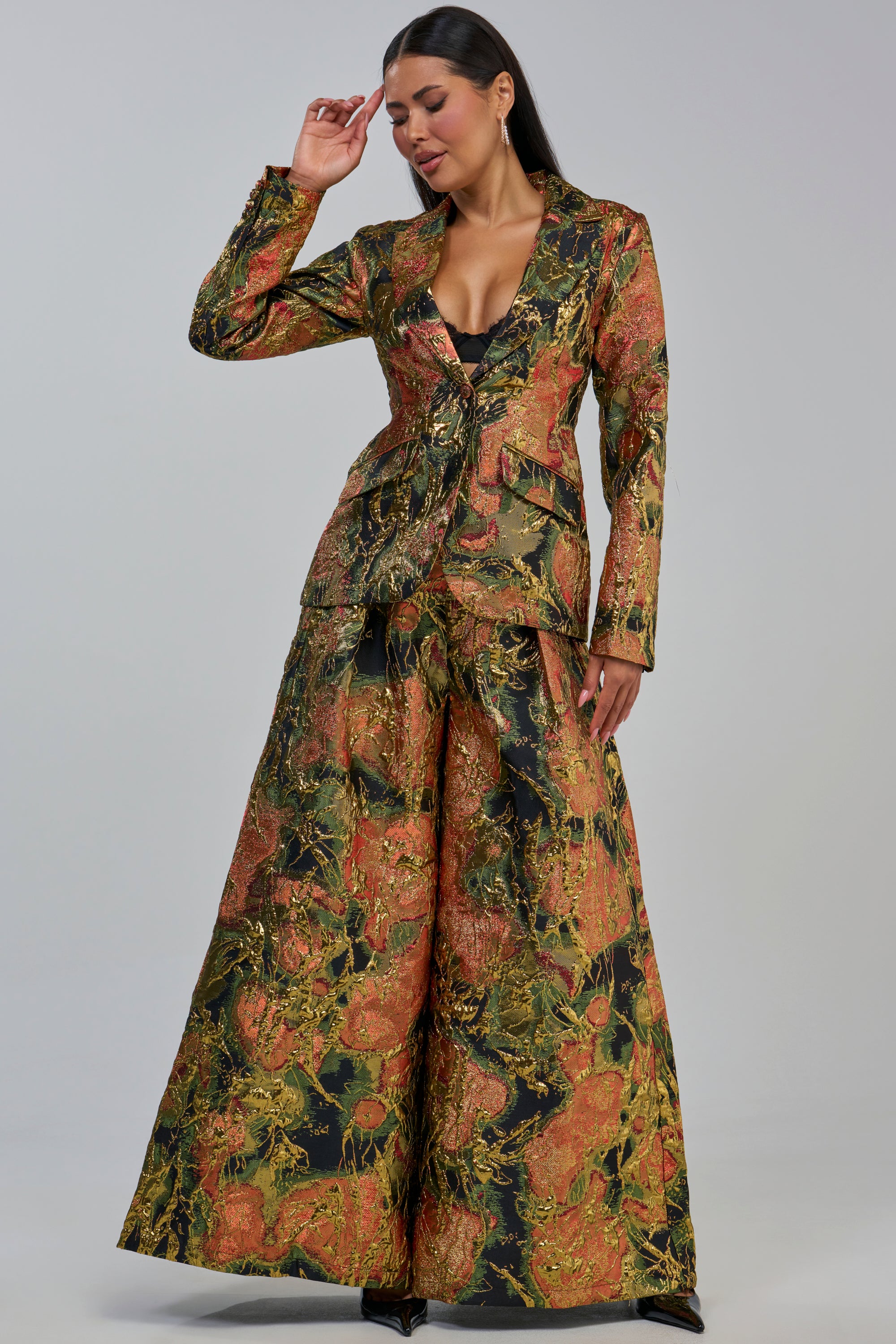 A woman wears the BUY MYSELF FLOWERS BROCADE TROUSER IN GOLD—a pair of wide-leg gold brocade trousers featuring orange and green tones—with a coordinating long blazer, posing with one hand near her head against a plain background.