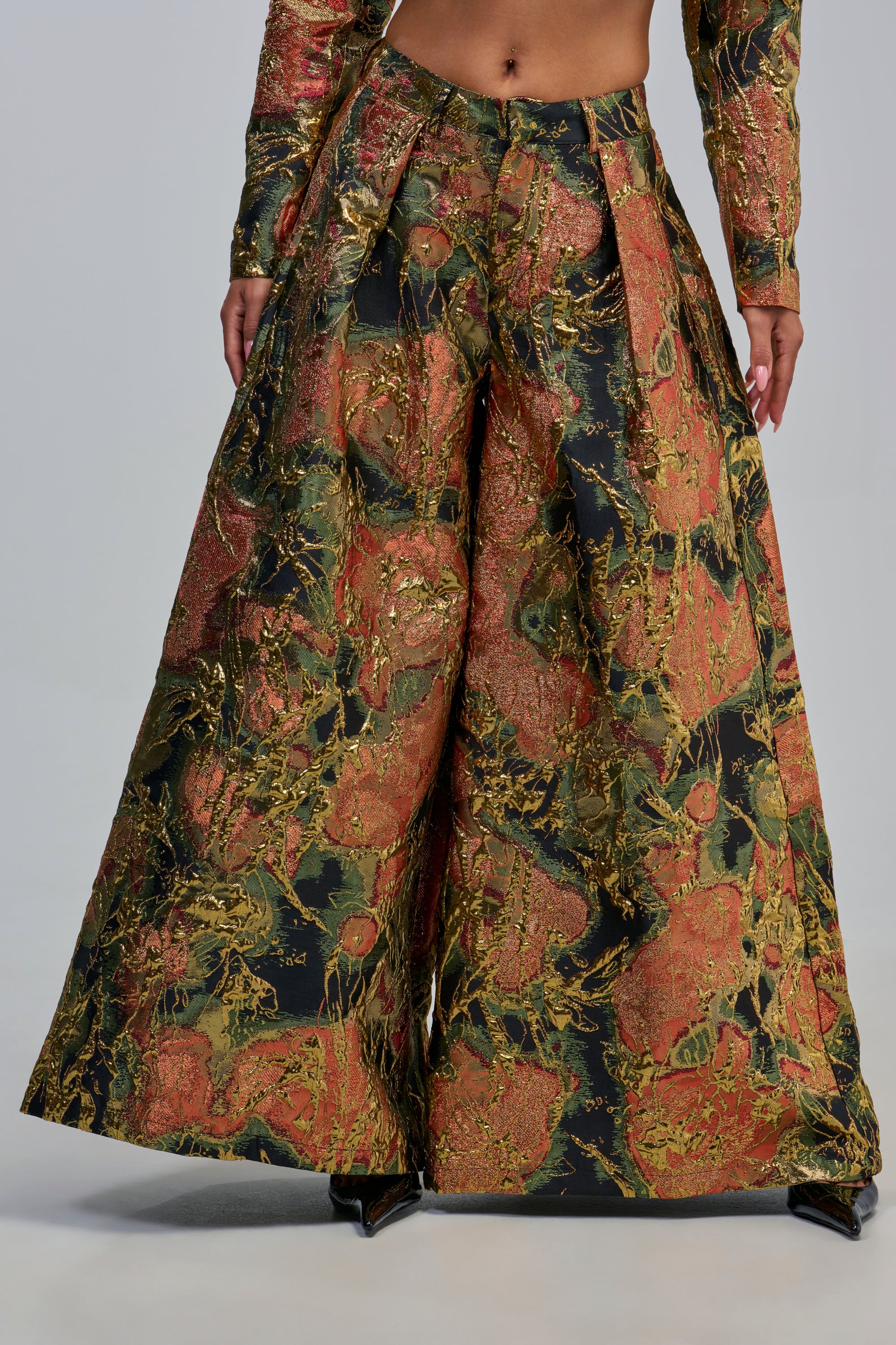 A person wearing the BUY MYSELF FLOWERS BROCADE TROUSER IN GOLD—wide-legged, high-waisted metallic brocade pants with a gold, red, and green abstract pattern—and pointed black shoes, shown from waist to feet.