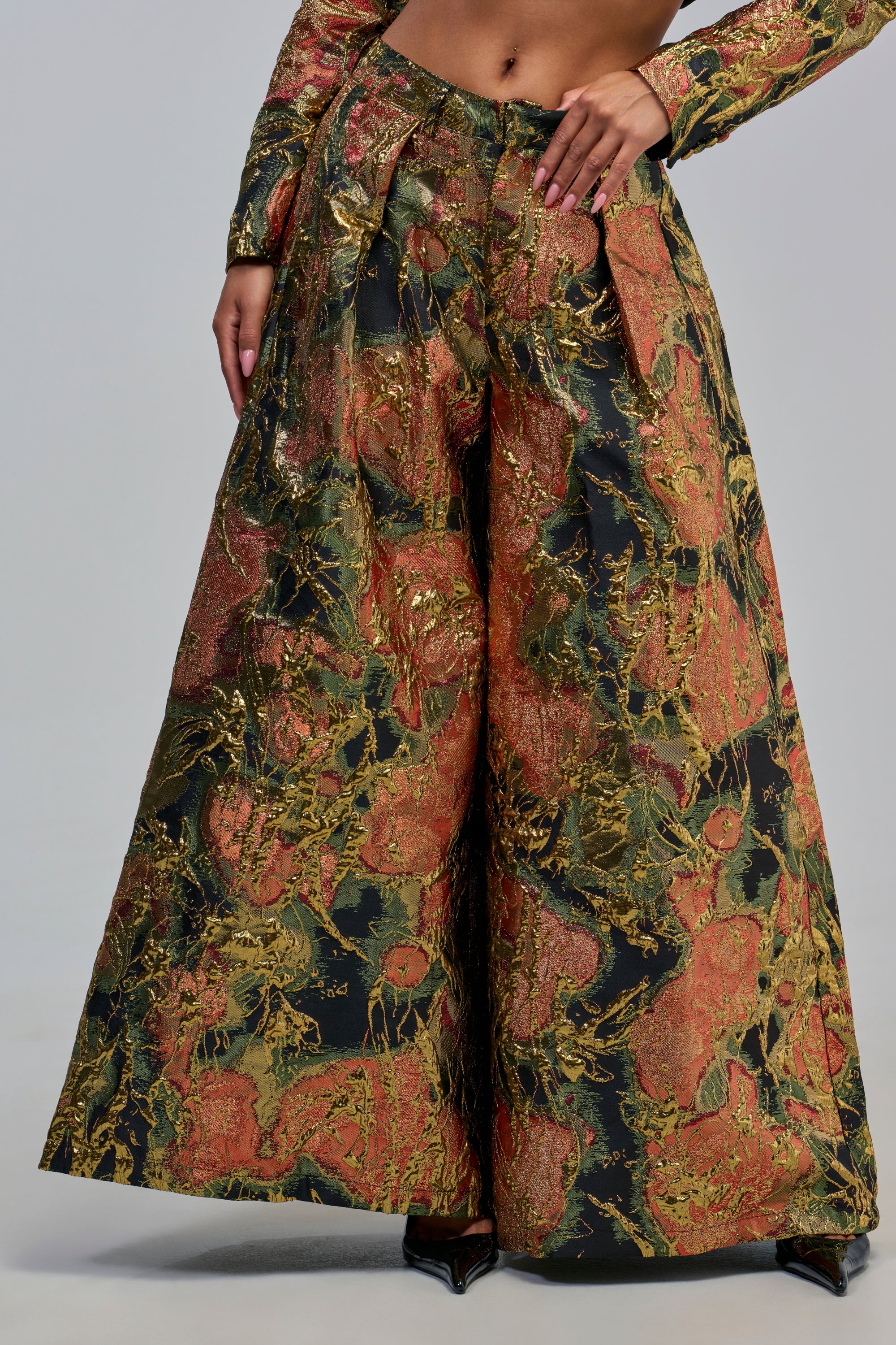 A person wearing the BUY MYSELF FLOWERS BROCADE TROUSER IN GOLD—high-waisted, wide-leg pants featuring a vibrant metallic gold, green, and orange abstract pattern—paired with a matching long-sleeve crop top for a bold evening look.