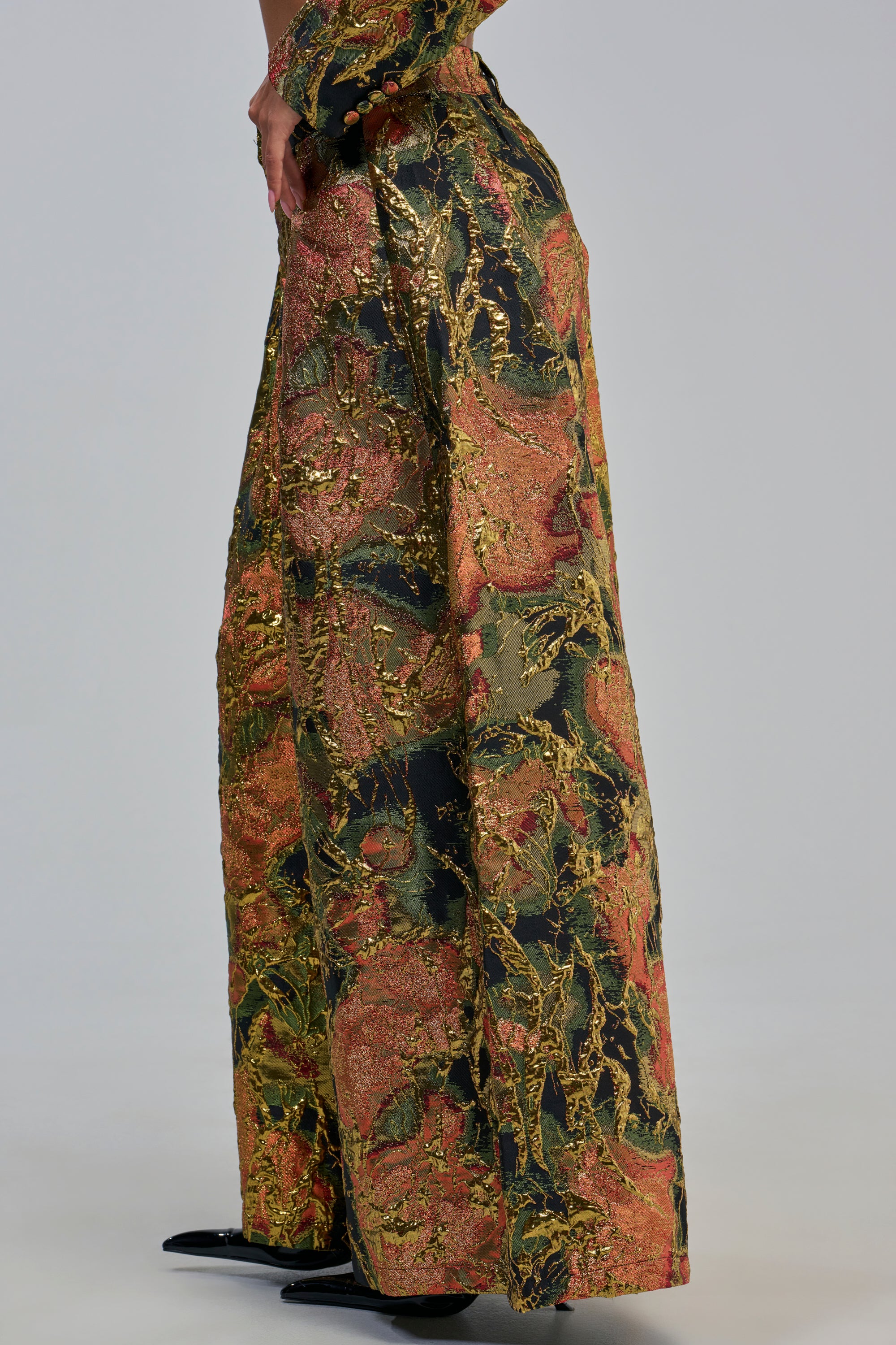 A person stands against a plain background wearing the BUY MYSELF FLOWERS BROCADE TROUSER IN GOLD, featuring a wide-leg silhouette and an abstract gold, orange, and green pattern.