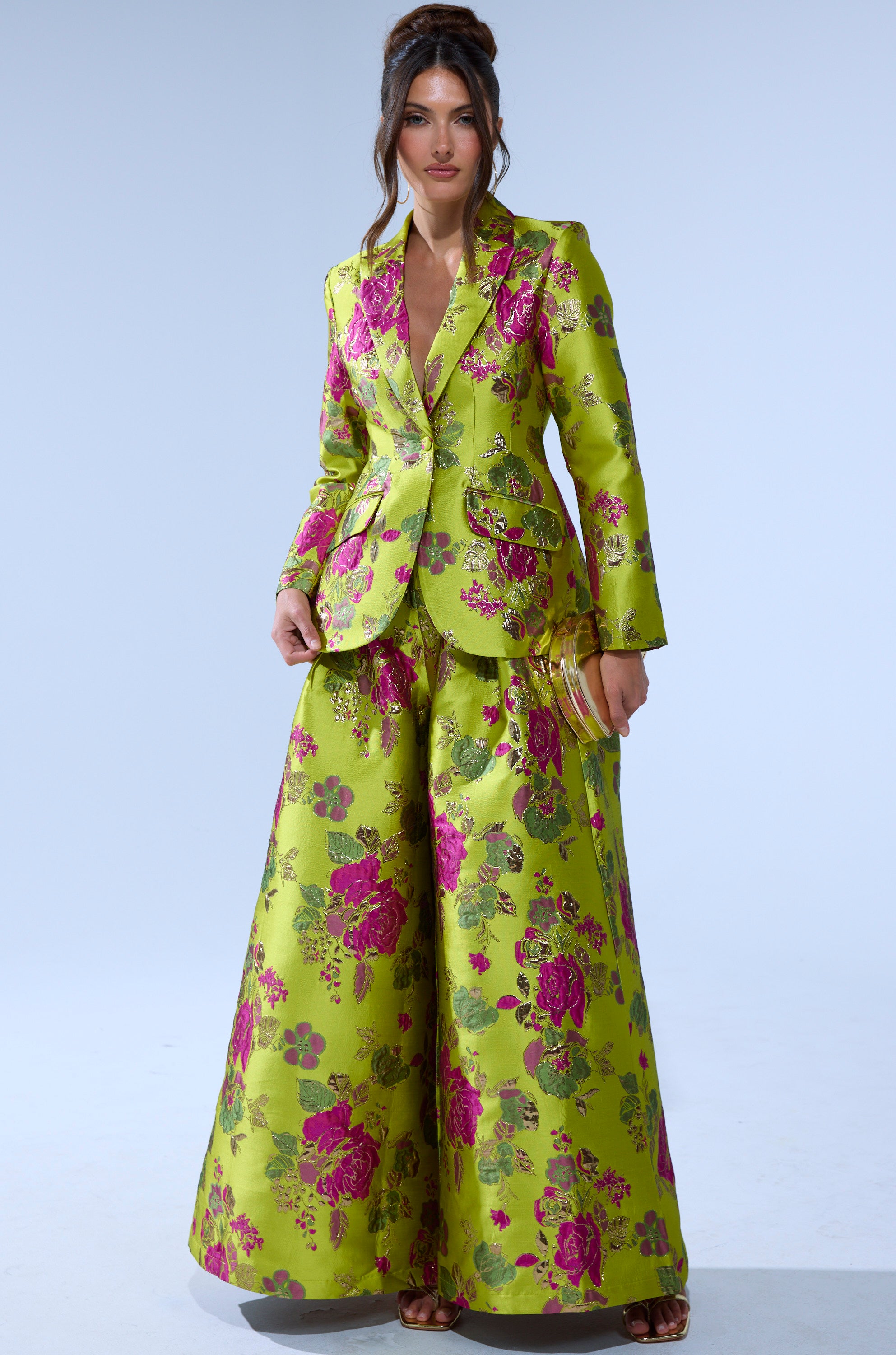 A woman wears the BUY MYSELF FLOWERS BROCADE TROUSER IN LIME—a bright green wide-leg trouser with pink and red floral print—paired with a matching blazer, a small clutch, and her hair styled in loose waves.