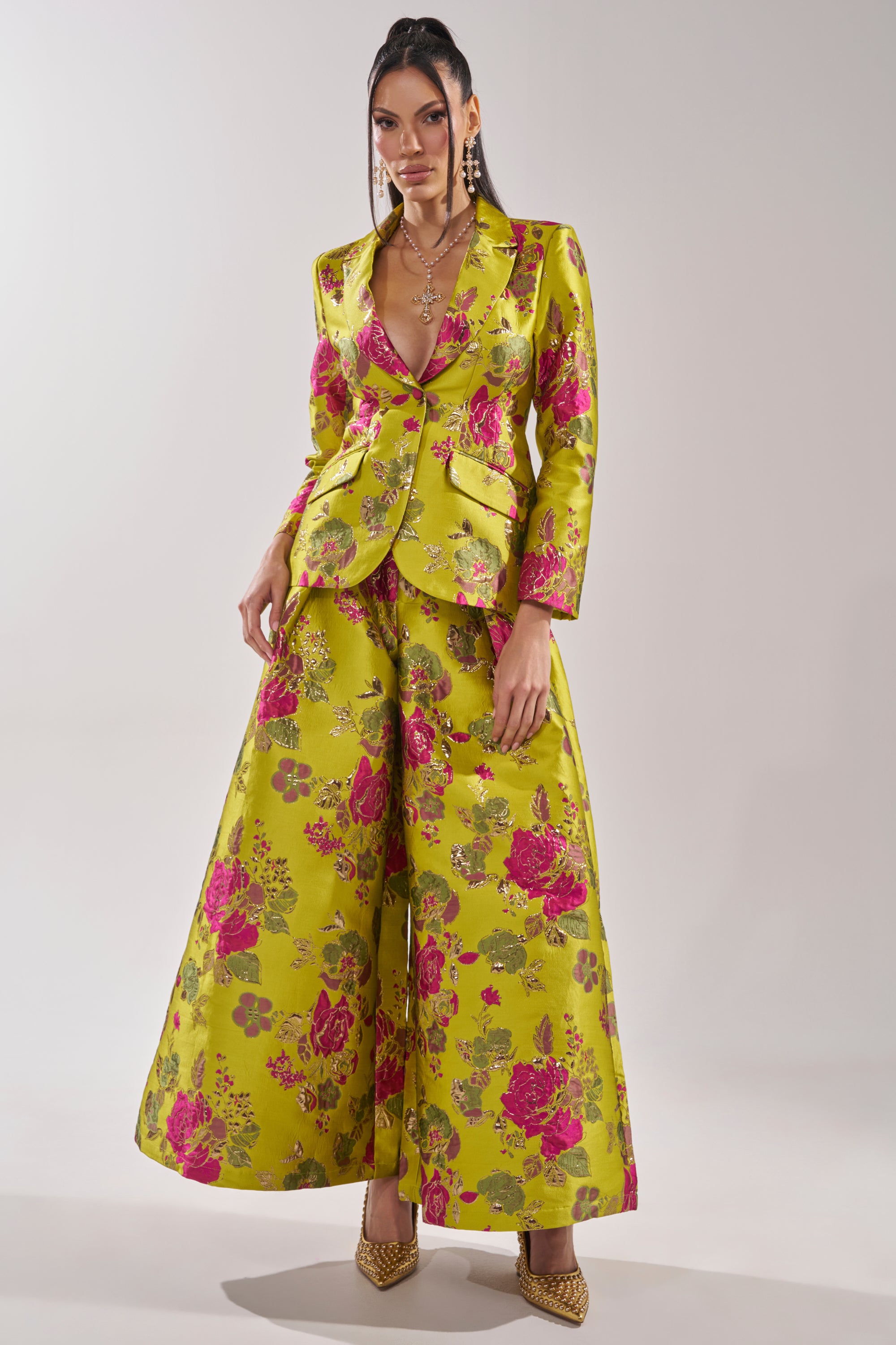 A woman poses in the BUY MYSELF FLOWERS BROCADE TROUSER IN LIME, featuring bright pink florals, styled with a matching blazer, layered necklaces, drop earrings, and pointed heels for a chic ensemble.