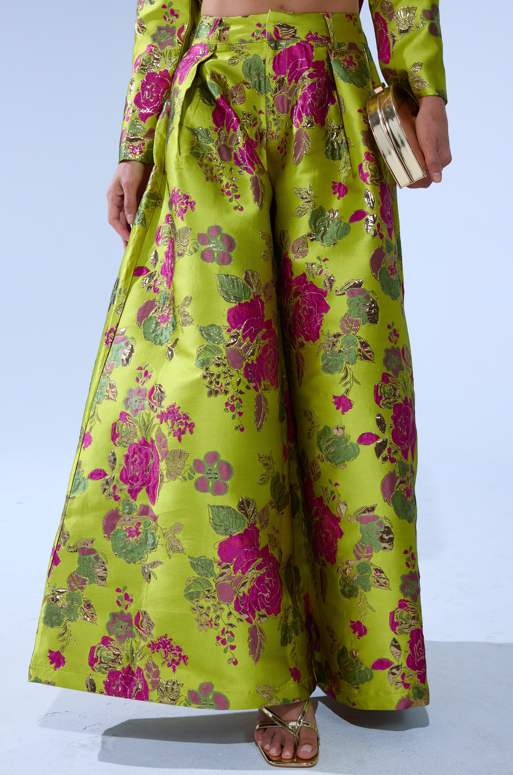 A person wears the BUY MYSELF FLOWERS BROCADE TROUSER IN LIME, a wide-leg style with bright green and pink florals, paired with gold sandals and a matching clutch. The photo is chest-up cropped.