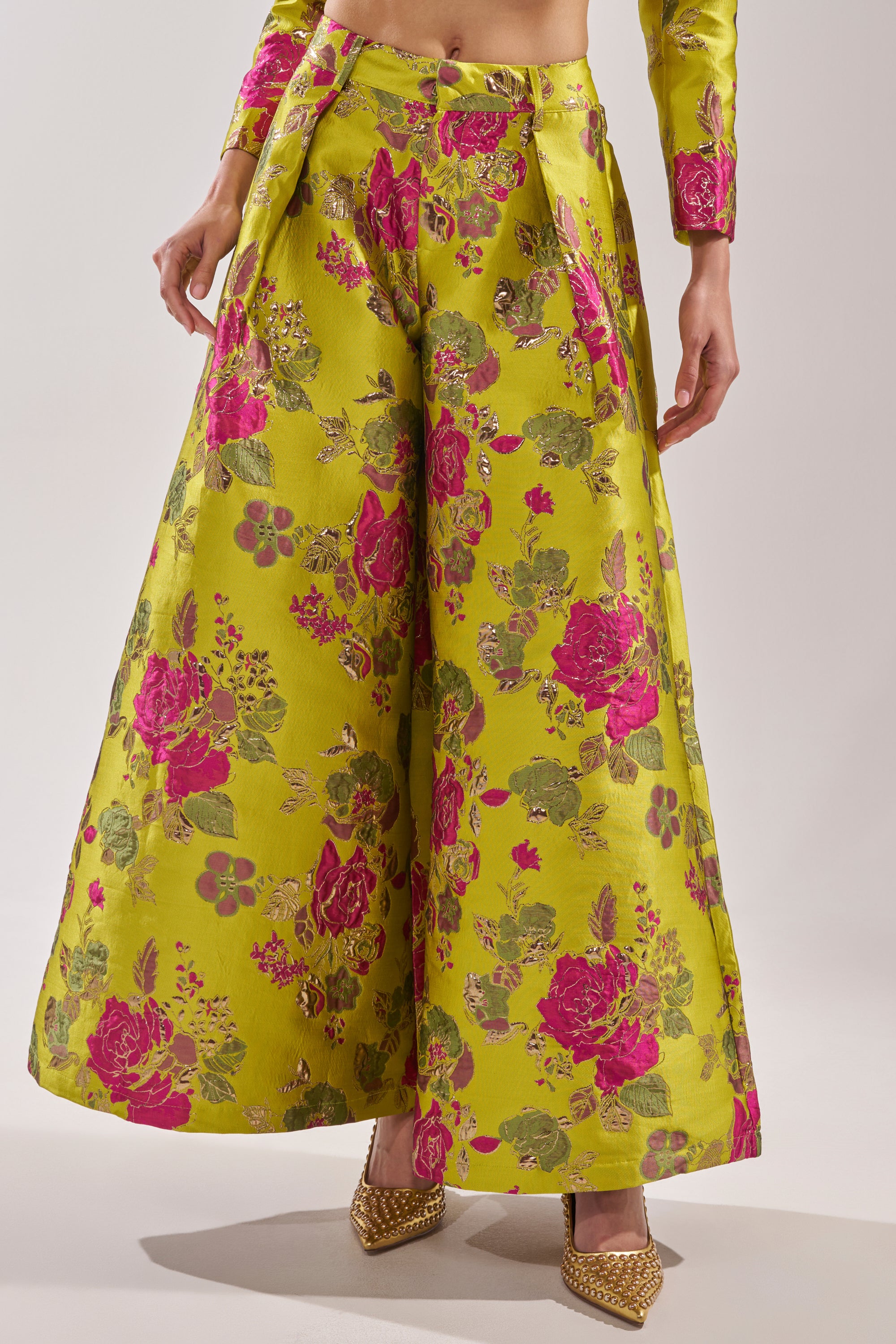 Someone wears the BUY MYSELF FLOWERS BROCADE TROUSER IN LIME—wide-leg, high-waisted trousers with a shiny pink and green floral pattern—paired with a matching cropped jacket and gold pointed-toe heels against a plain light gray background.