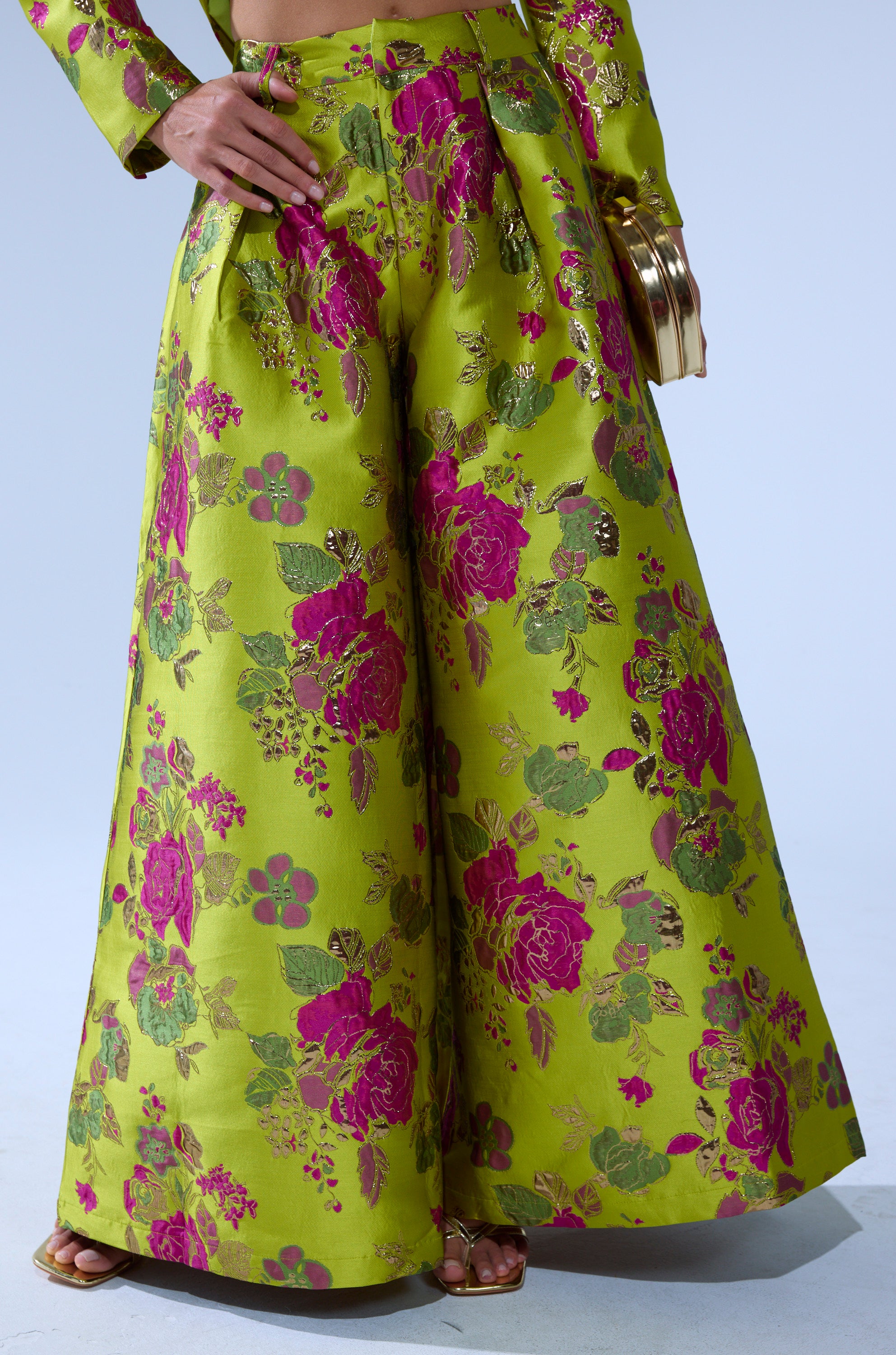 A person wears the BUY MYSELF FLOWERS BROCADE TROUSER IN LIME, featuring a wide-leg silhouette and bold pink and gold floral print on lime green, paired with gold sandals and clutch. Only the lower half of the body is shown.