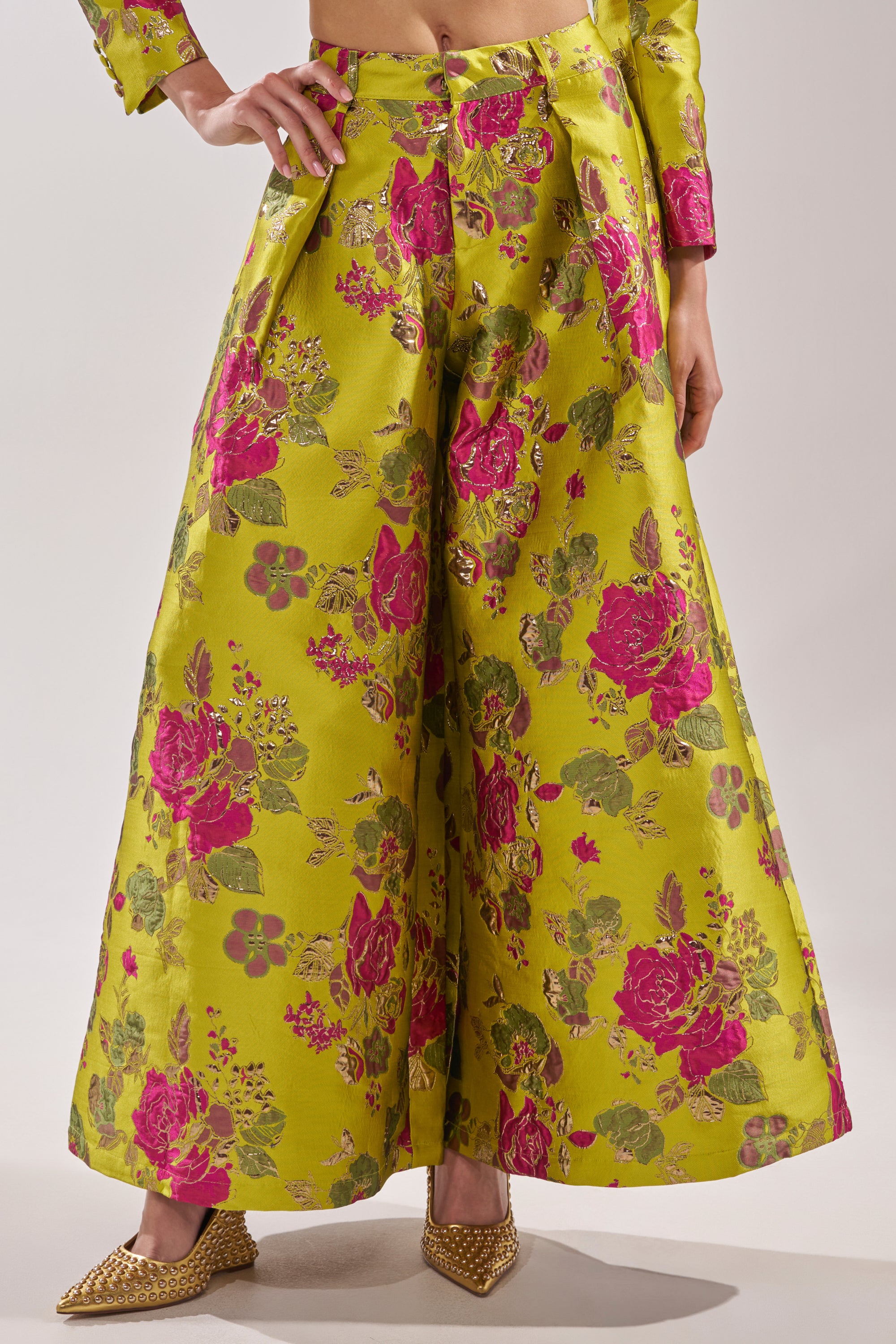 A person wears the BUY MYSELF FLOWERS BROCADE TROUSER IN LIME, featuring a vibrant yellow-green base and bold pink floral pattern, styled with a matching cropped jacket and pointed gold shoes. Their face is not visible.