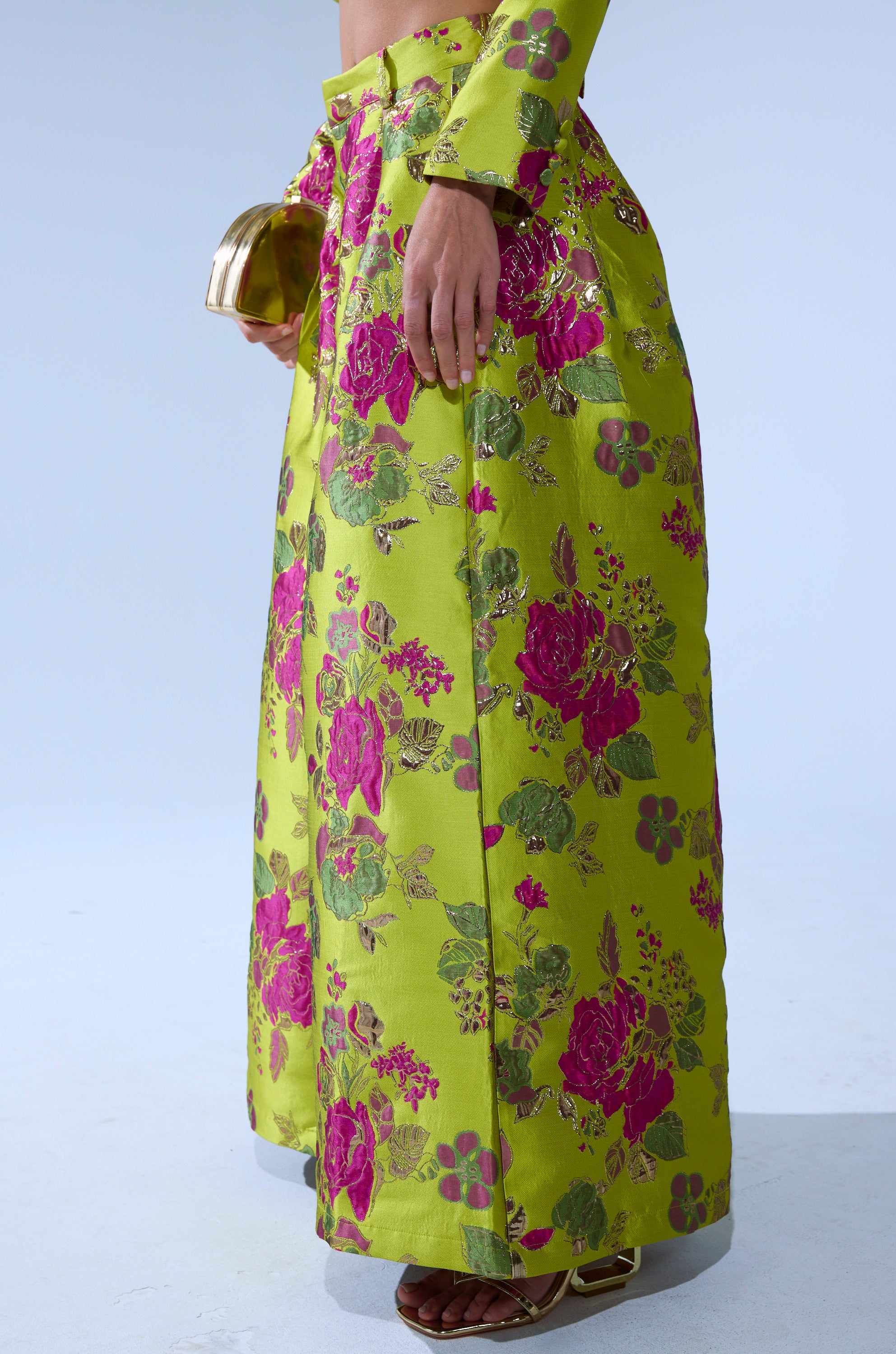 Someone wears the BUY MYSELF FLOWERS BROCADE TROUSER IN LIME, featuring metallic pink and gold floral accents, paired with a gold clutch and brown heeled sandals.