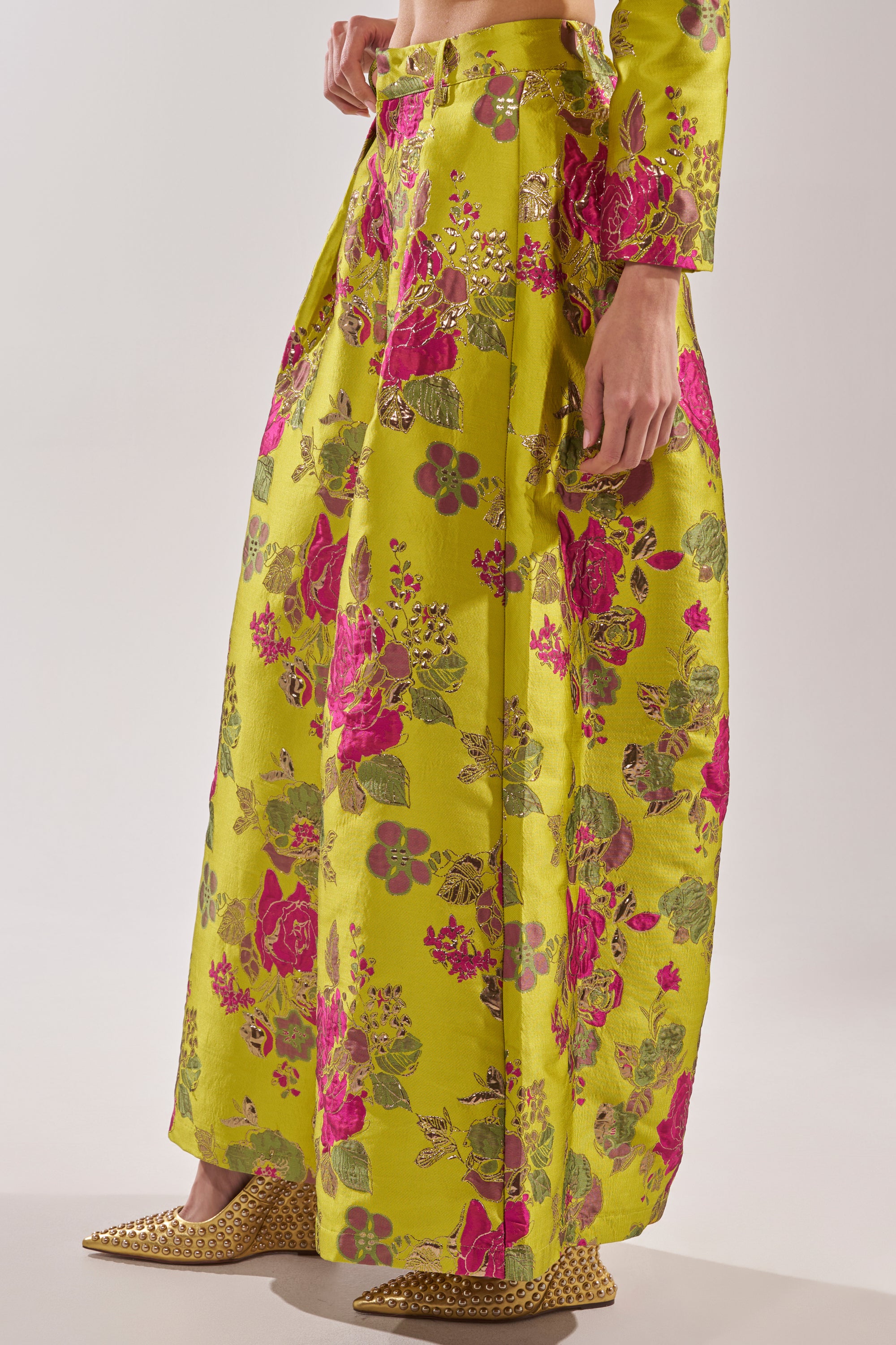 The look features the BUY MYSELF FLOWERS BROCADE TROUSER IN LIME—high-waisted, wide-leg trousers with pink floral and metallic details. Paired with pointed gold studded shoes, only the lower body is shown.