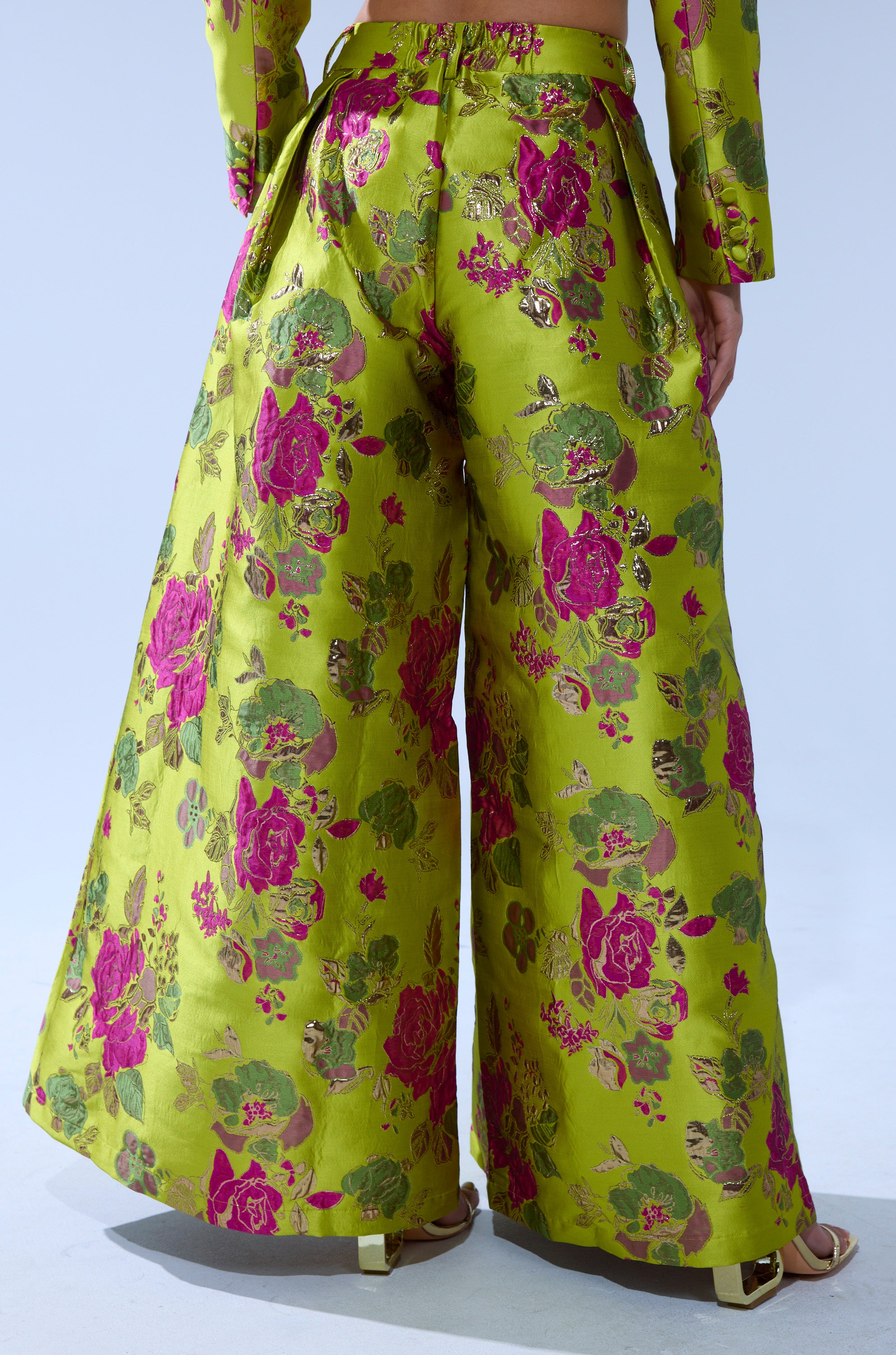 Wearing the BUY MYSELF FLOWERS BROCADE TROUSER IN LIME, featuring shiny green fabric with a pink and gold floral pattern, and matching sandals. The photo is taken from behind, highlighting the standout pants.