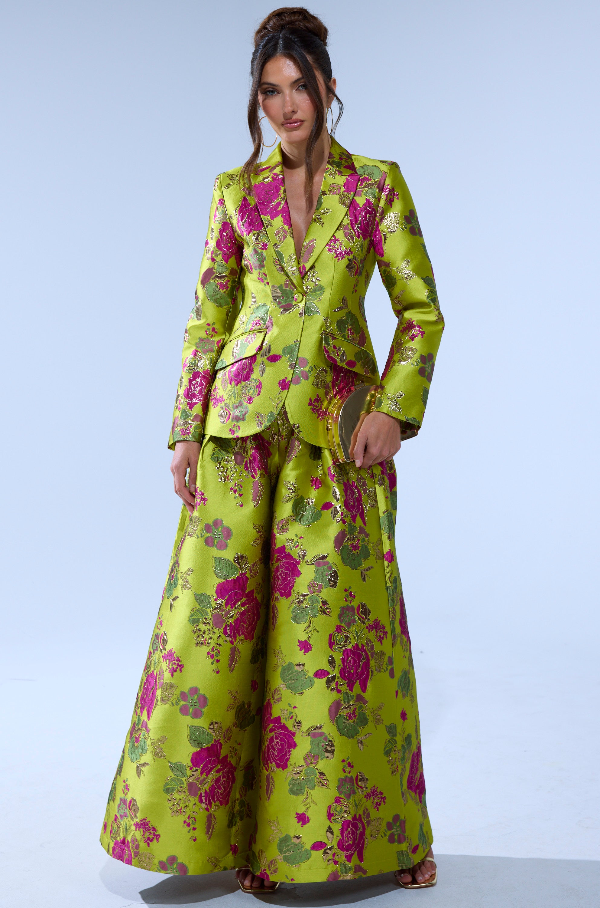 A woman wears the BUY MYSELF FLOWERS BROCADE TROUSER IN LIME, featuring a shiny lime green hue and pink-gold floral pattern, paired with a matching fitted jacket. Her hair is styled in a loose updo against a plain light background.