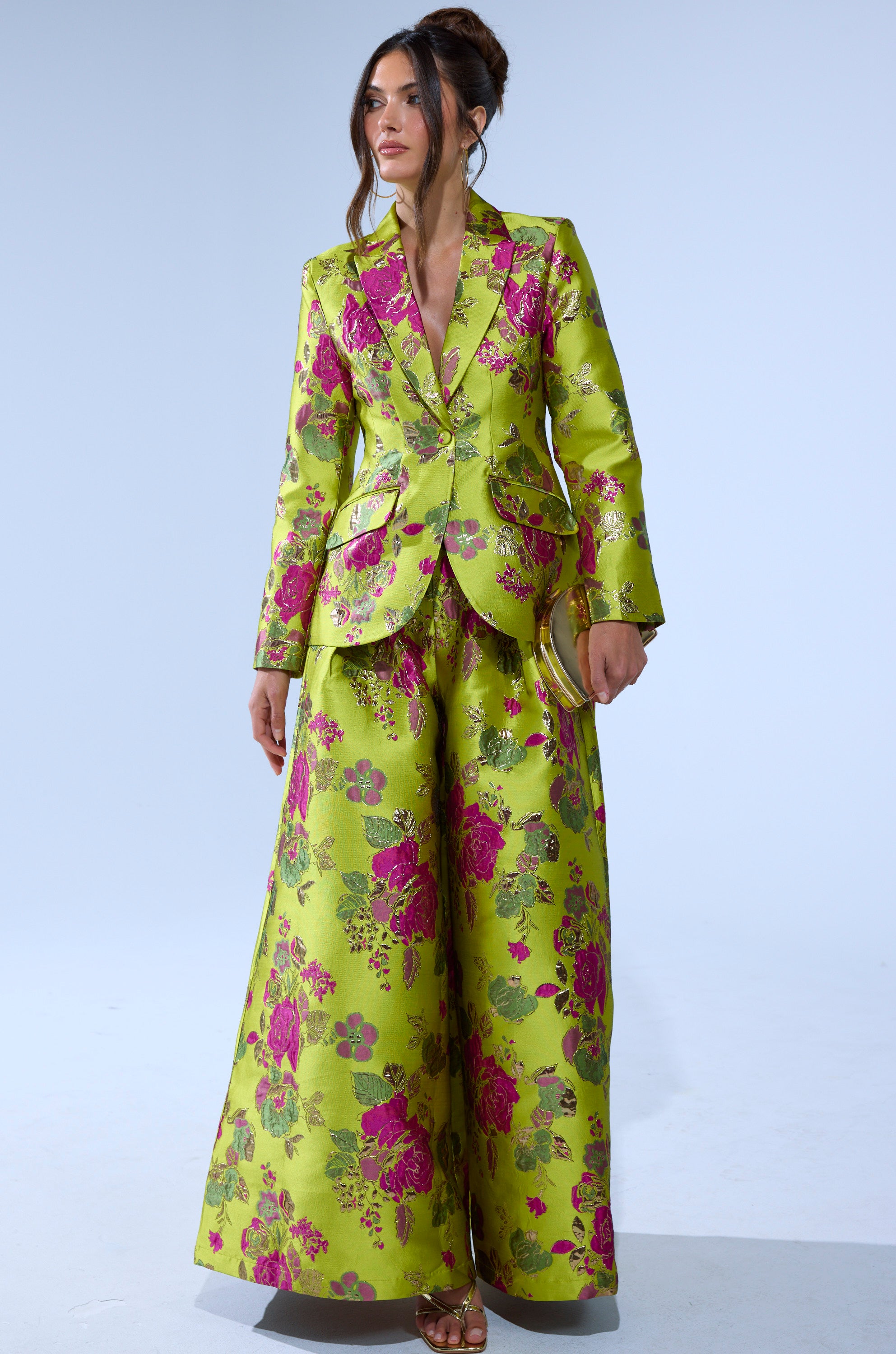 A woman wears the BUY MYSELF FLOWERS BROCADE TROUSER IN LIME, a vibrant wide-leg pant with a bold pink floral print, paired with a matching blazer. Her long brown hair is styled up and she completes the look with open-toed heels.
