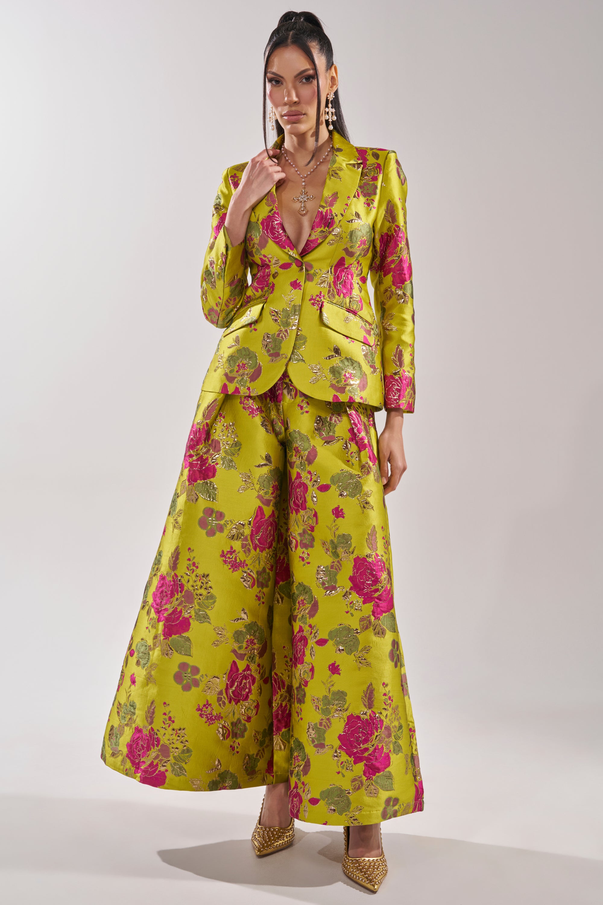 A woman poses confidently in the BUY MYSELF FLOWERS BROCADE TROUSER IN LIME, a bright yellow-green suit with a pink floral pattern, styled with gold heels and layered jewelry in a studio setting.