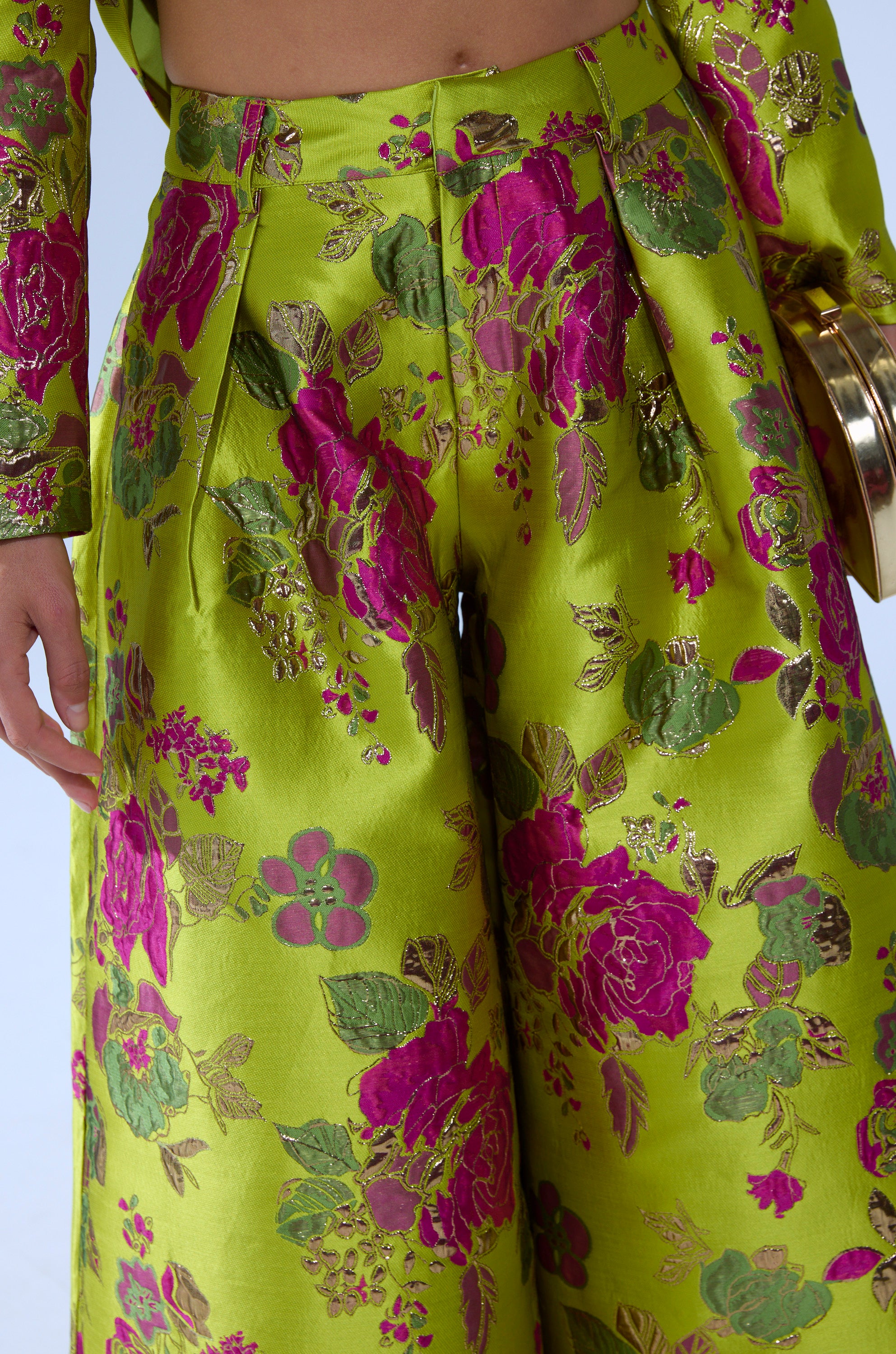 A close-up of a person wearing the BUY MYSELF FLOWERS BROCADE TROUSER IN LIME, featuring shiny lime green fabric with bold pink and gold floral patterns, paired with a matching jacket and holding a round gold clutch.
