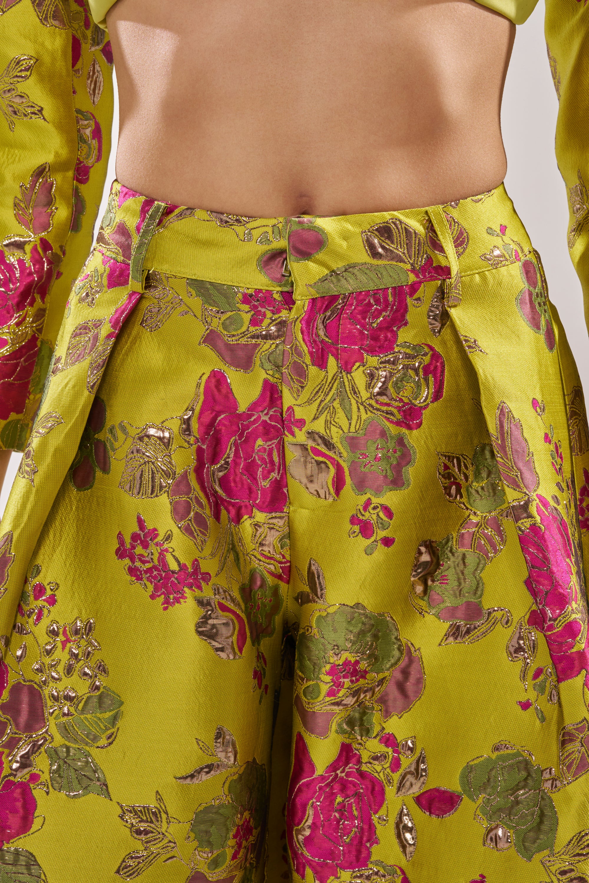 A close-up shows a person in the BUY MYSELF FLOWERS BROCADE TROUSER IN LIME, featuring a high-waisted, wide-leg fit and vibrant floral pattern in pink, green, and gold on shiny lime fabric. Their bare midriff is partly visible above the waistband.