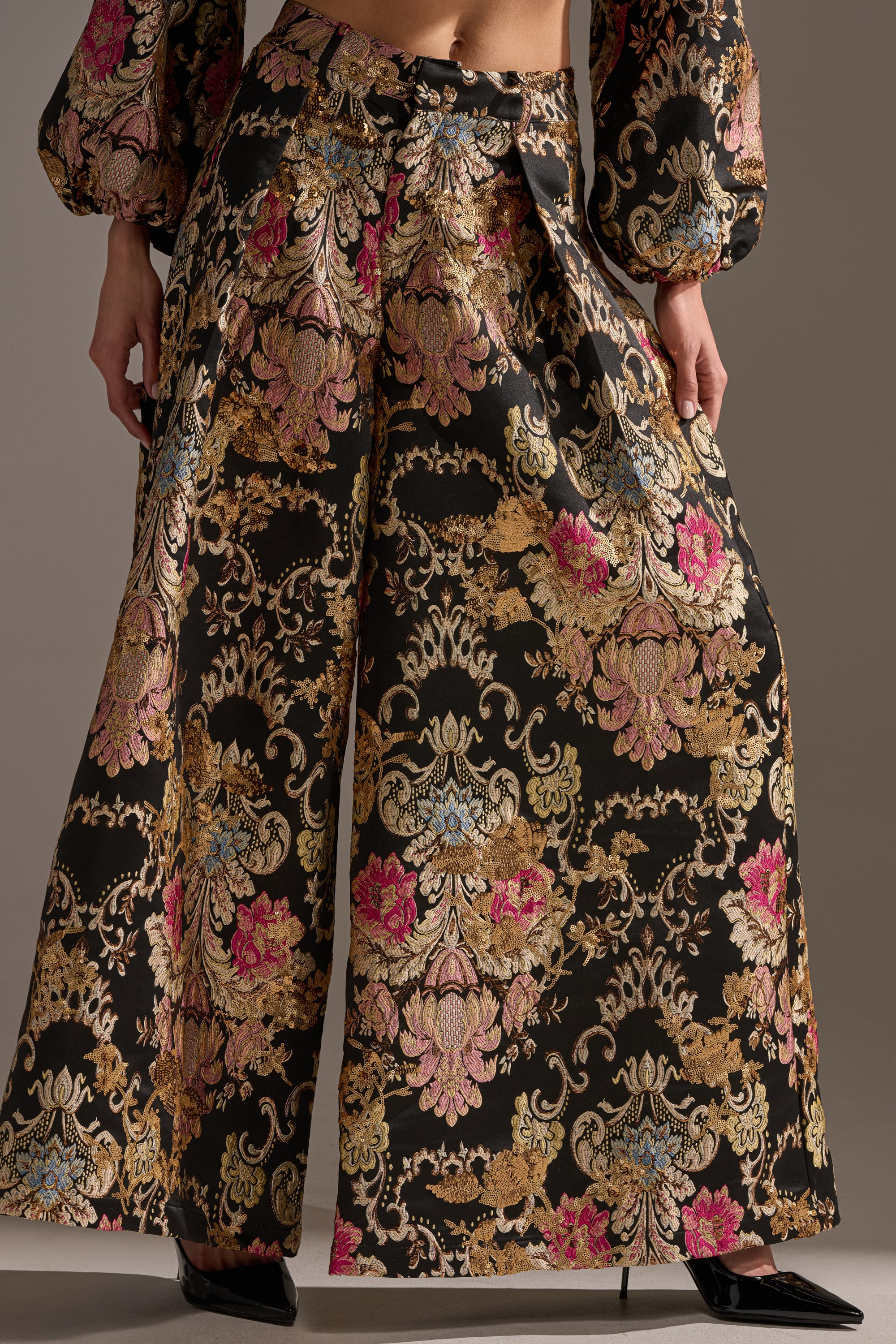 Wearing the LUXE ENERGY PALAZZO PANT in vibrant floral brocade—gold, pink, and blue on black—with a matching long-sleeve top and black pointed-toe heels.