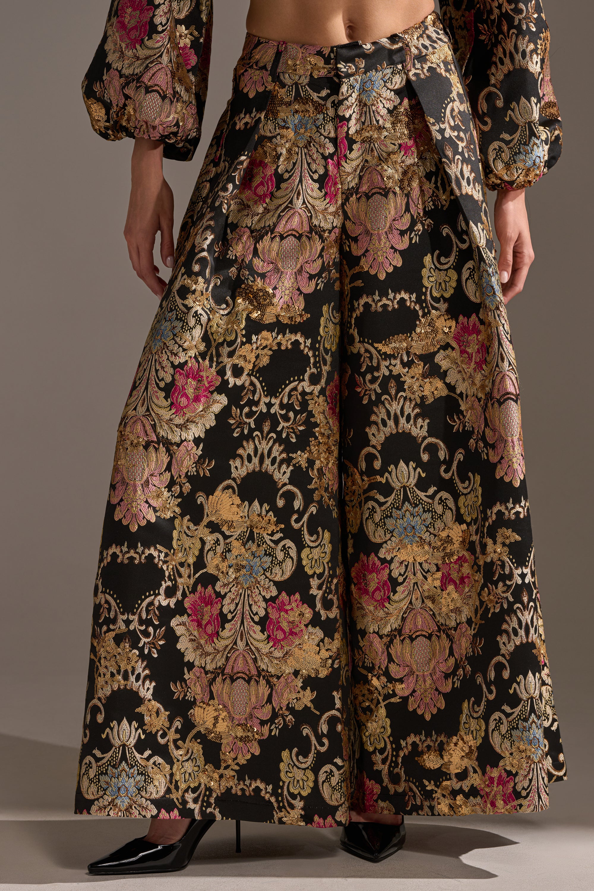 Wearing the LUXE ENERGY PALAZZO PANT with a wide-leg silhouette and ornate baroque floral brocade in gold, pink, and blue, paired with a matching cropped top and black pointed-toe heels.